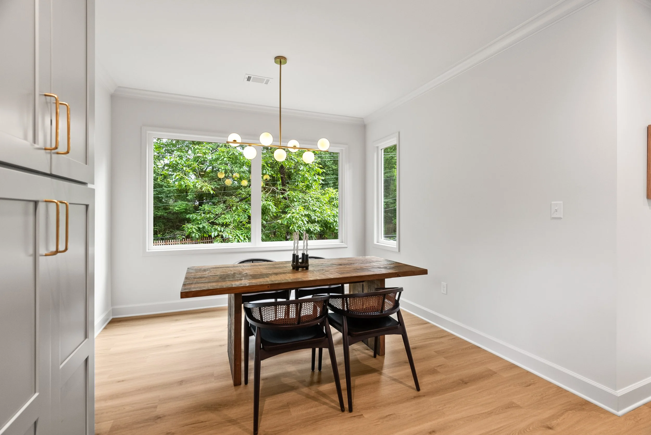 A dining area with a wooden table, black chairs, and a chandelier with globe bulbs hanging from the ceiling. There are three large windows providing a view of greenery outside.