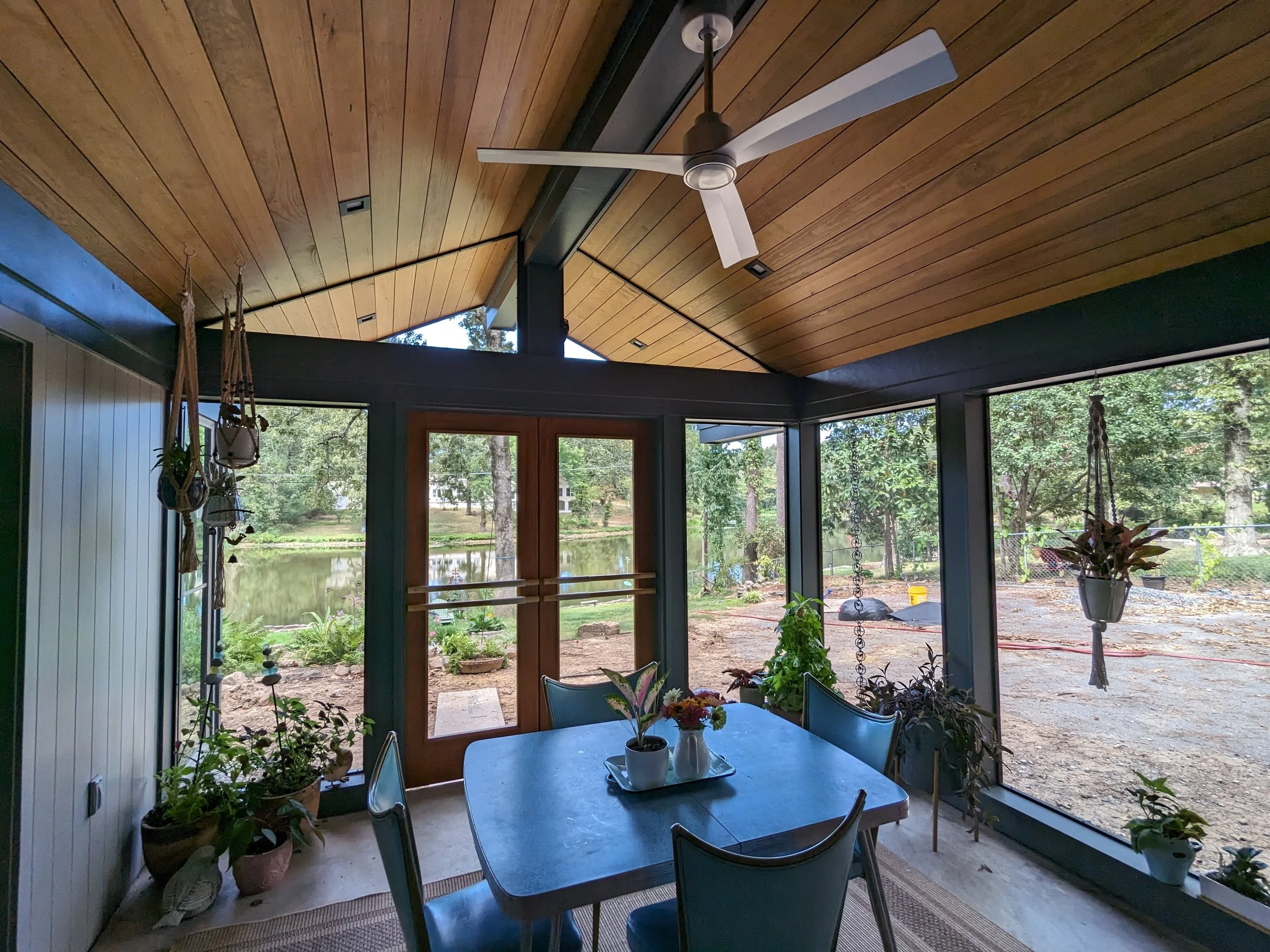 A sunlit screened porch with a wooden ceiling and ceiling fan, overlooking a backyard with trees, a pond, and a gravel area. There are potted plants and hanging decorations inside, and a blue table with a vase of flowers in the foreground.