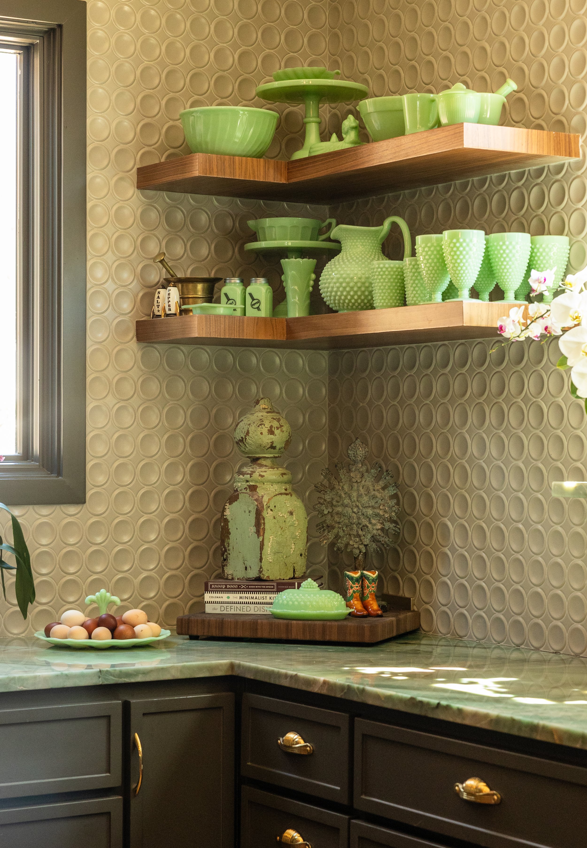 Kitchen with black cabinets and gold handles, green marble countertop, and floating wooden shelves holding green dishware and decor. Patterned wallpaper in the background and decorative items on the counter including eggs, books, and vintage bottles.