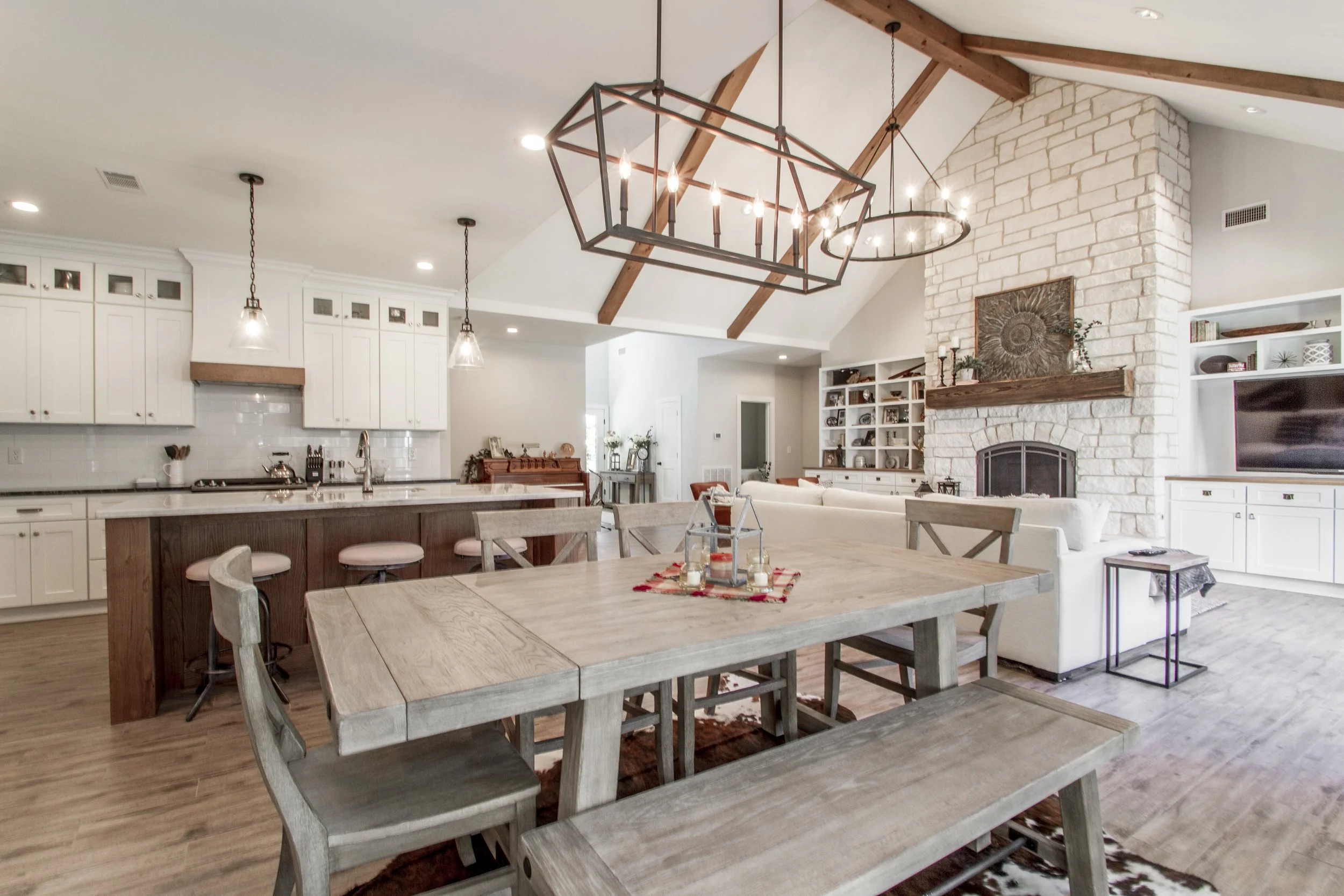 Open-concept kitchen and living room with white cabinetry, a brick fireplace, wooden beams, and a dining table with chairs.