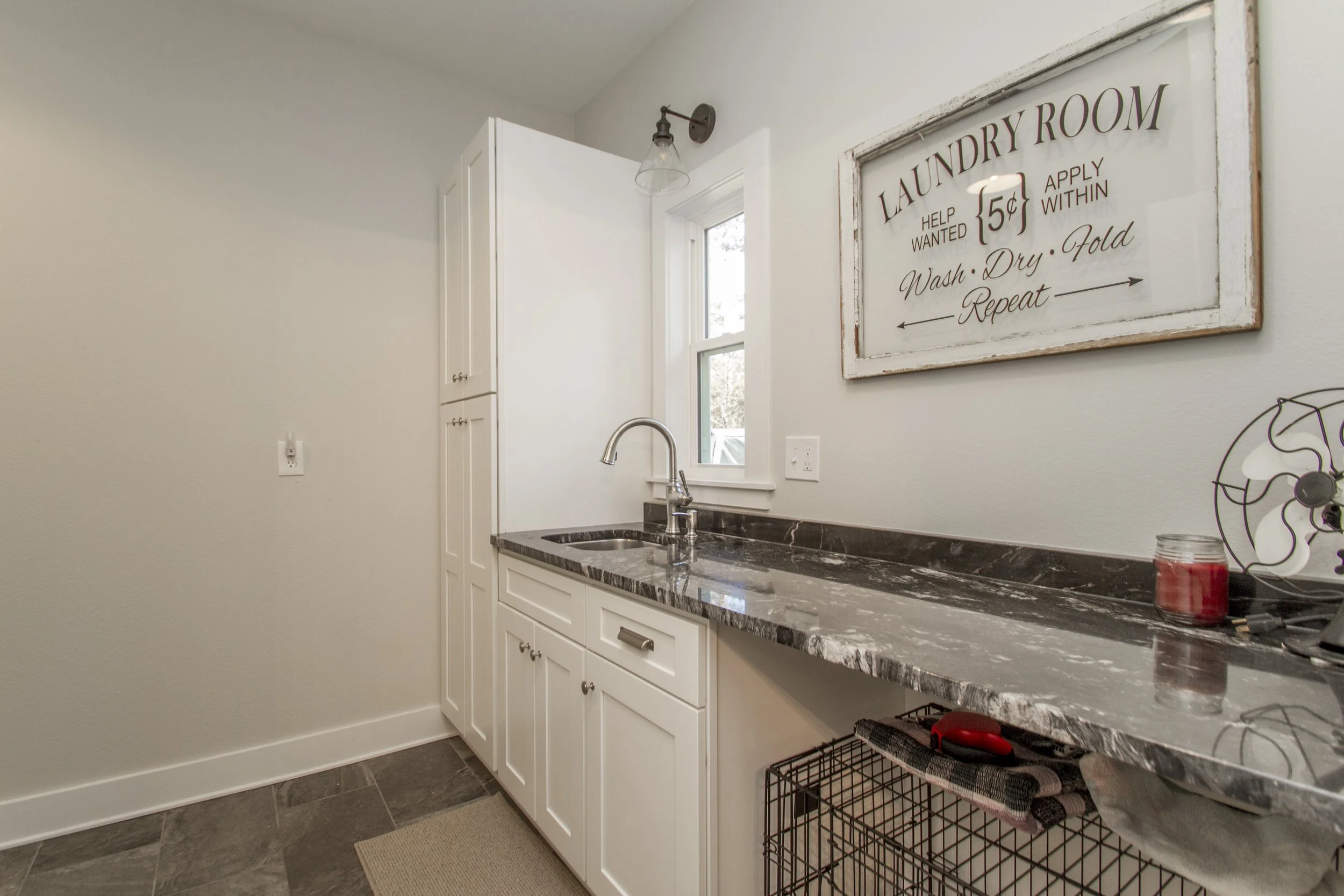 A laundry room with white cabinets, a black marble countertop, and a stainless steel sink. There is a window above the sink and a basket below the counter with folded towels. A sign on the wall reads 'Laundry Room' with instructions and a fee for hel