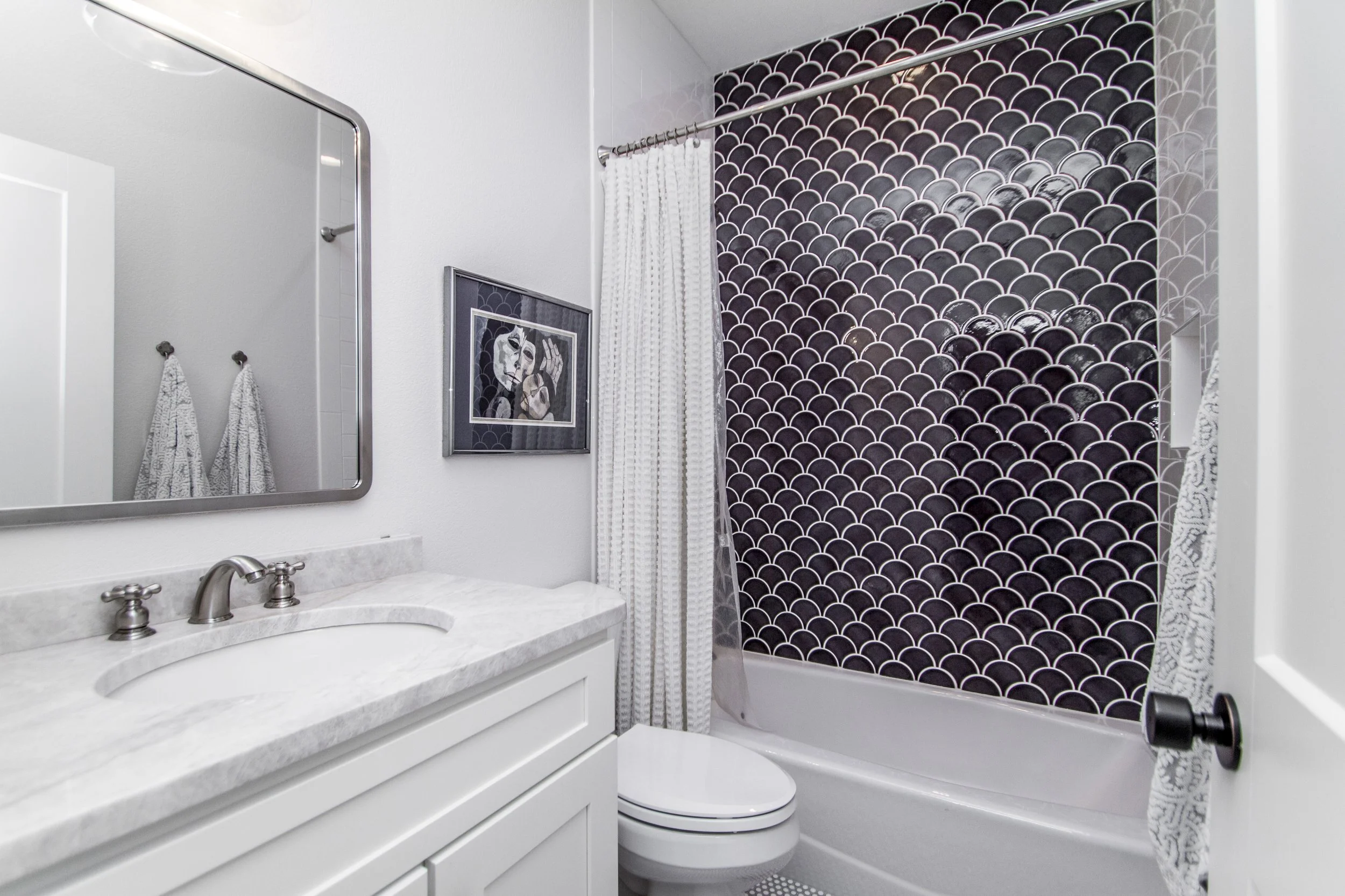 A bathroom with a white vanity sink, marble countertop, and a mirror. Towel hooks with patterned towels, framed artwork, and a shower with black scalloped tile. White shower curtain partially covers the tub.
