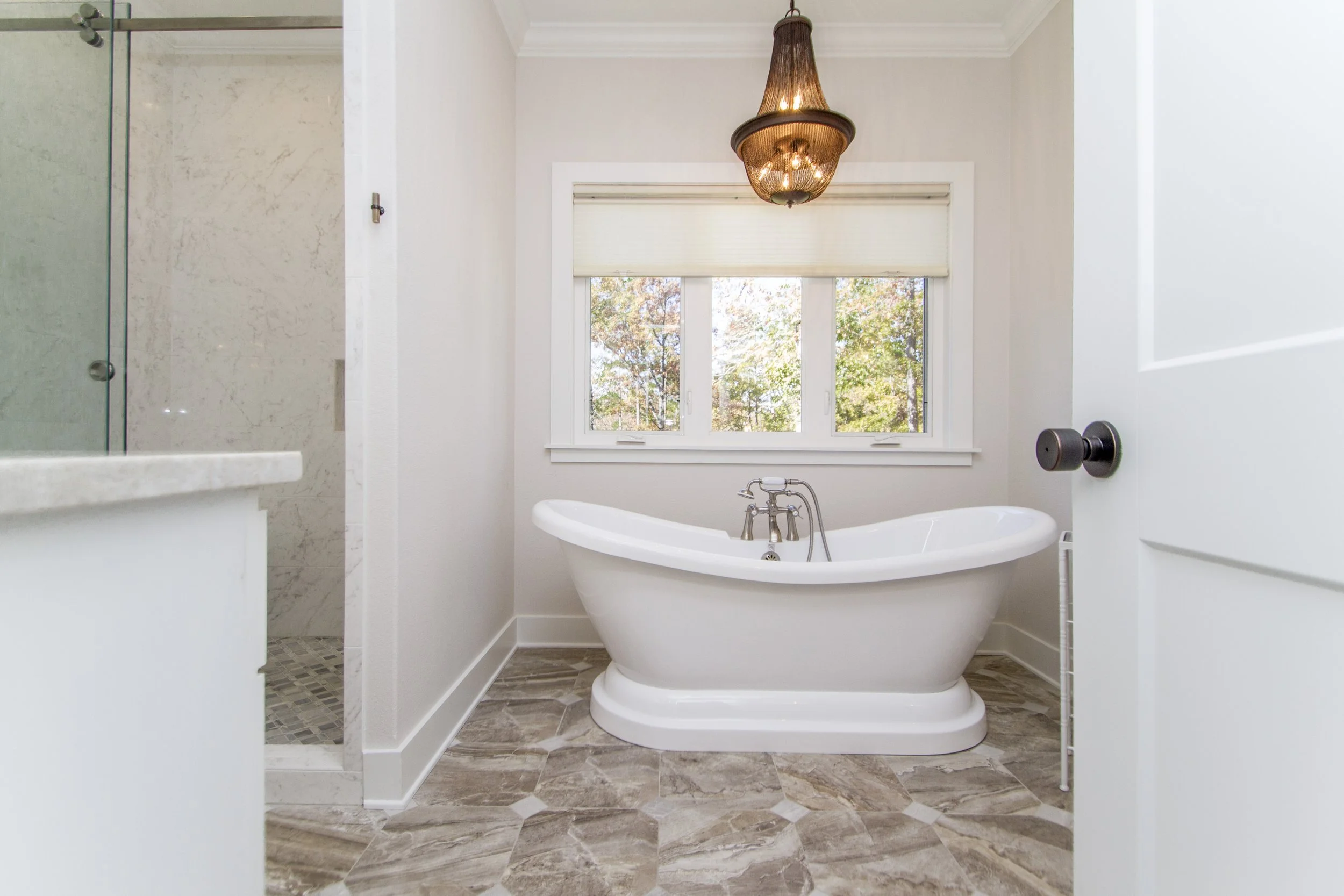 A bathroom with a clawfoot bathtub under a window, a chandelier hanging from the ceiling, and a tiled floor.