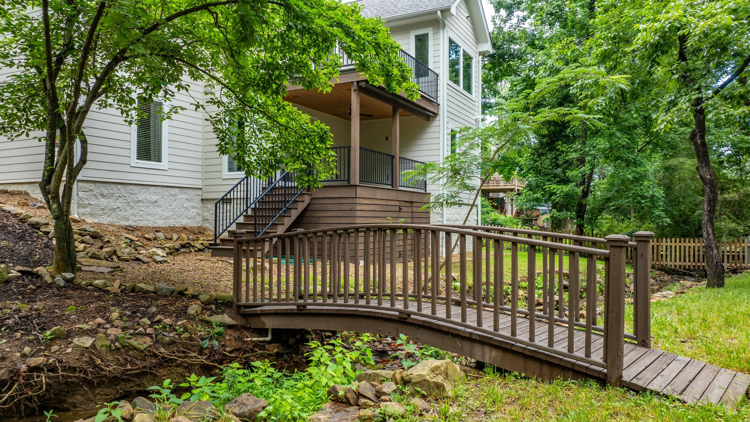 A two-story house with white siding, a deck with stairs, and a small bridge crossing over a rocky creek, surrounded by green trees and lawn.