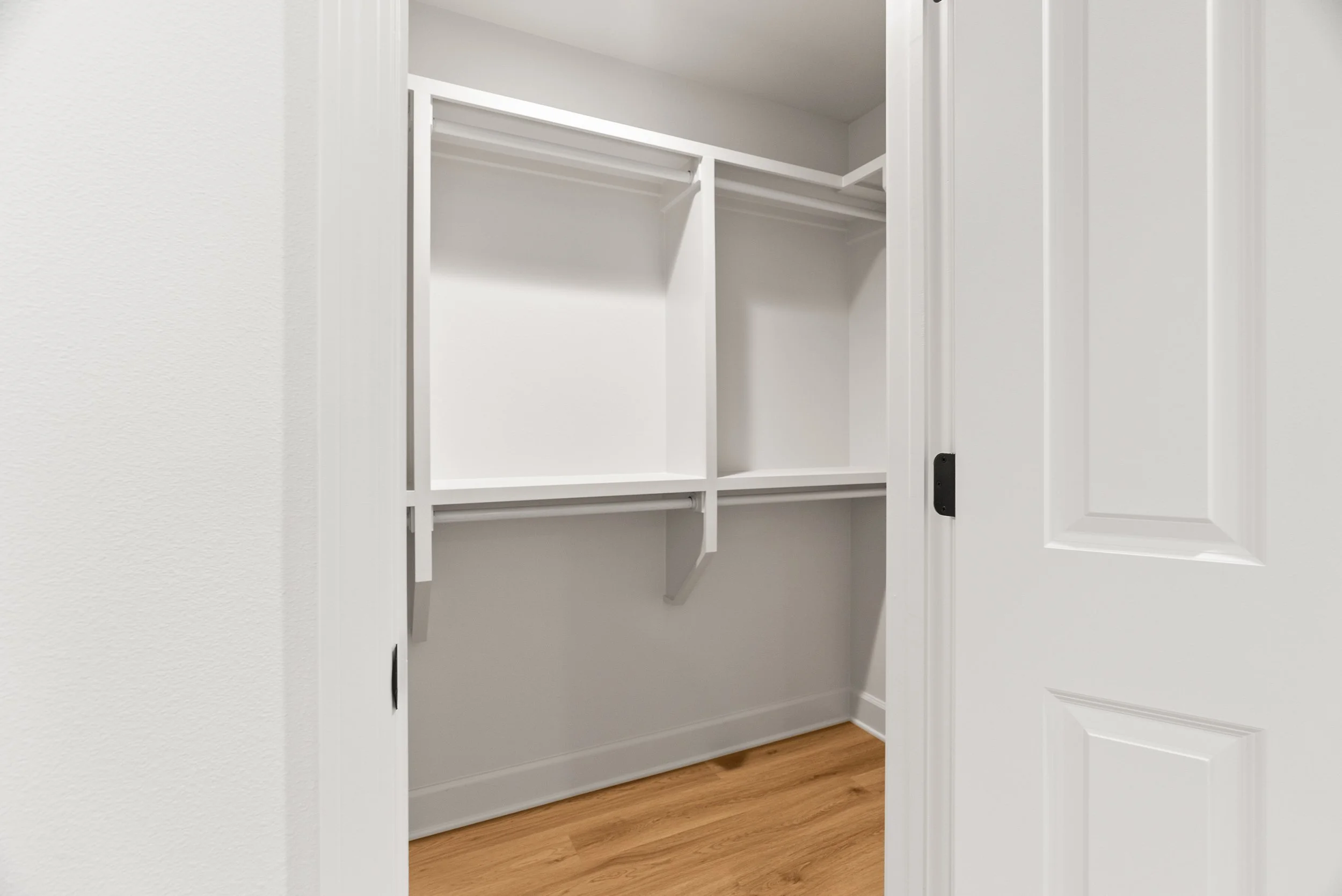 An empty white closet with wires and shelves, partially open door, white walls, and brown hardwood flooring.