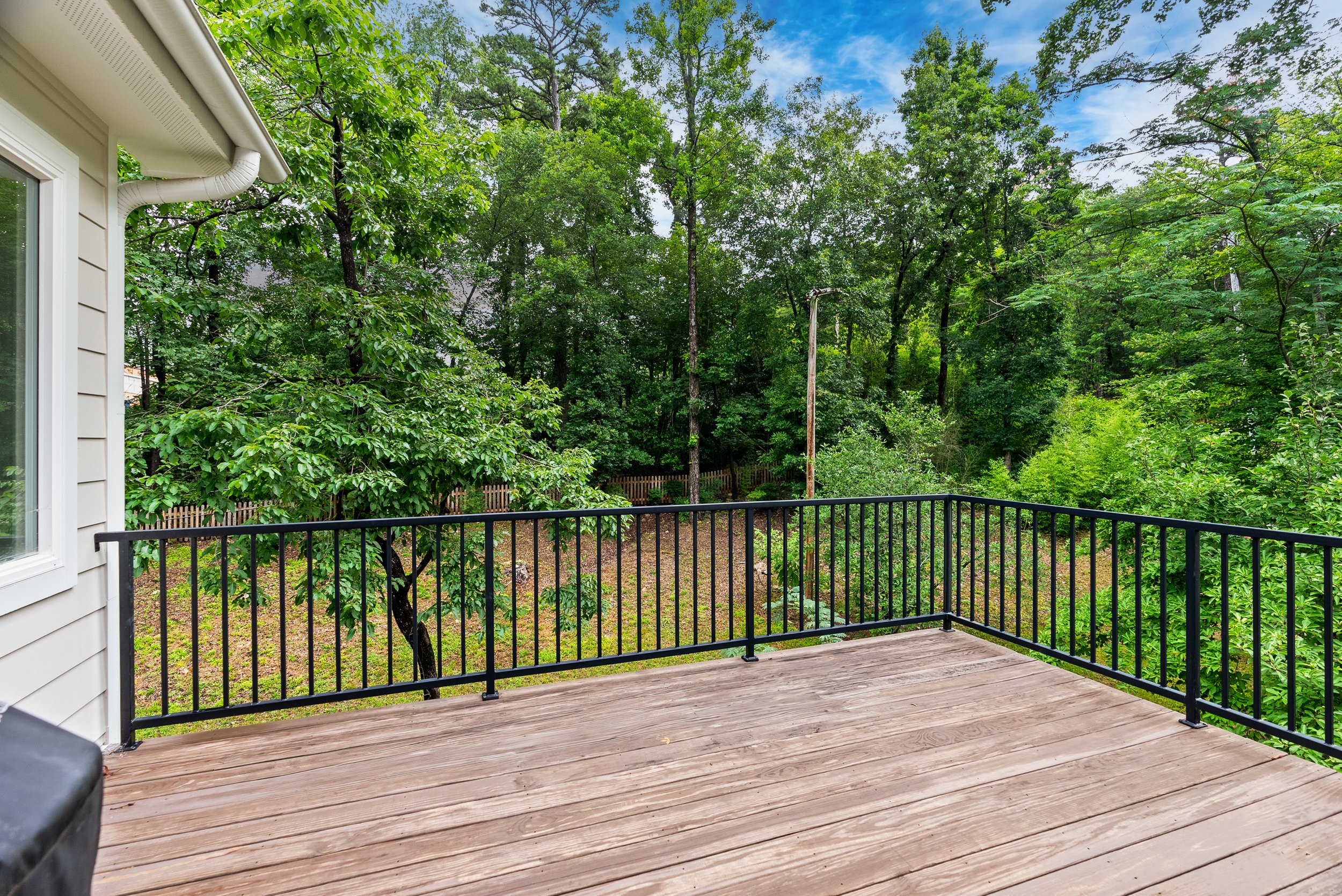 A wooden deck with black metal railing overlooks a lush green backyard with trees and bushes. The edge of the house with beige siding and a white window is visible on the left. The sky is partly cloudy with some blue showing through.