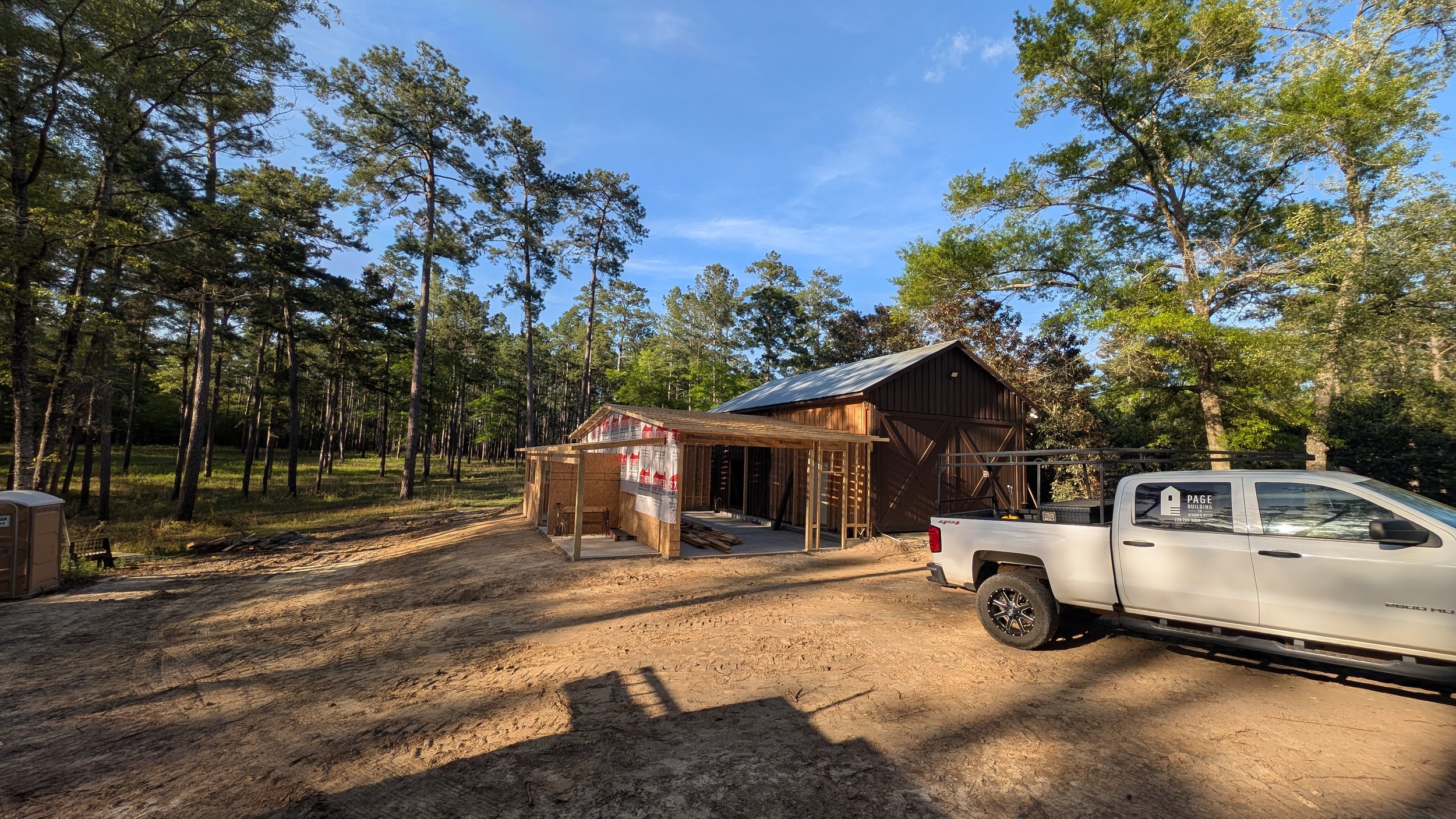 Construction site with a partially built wooden structure, white pickup truck parked nearby, wooded area in the background, clear blue sky.