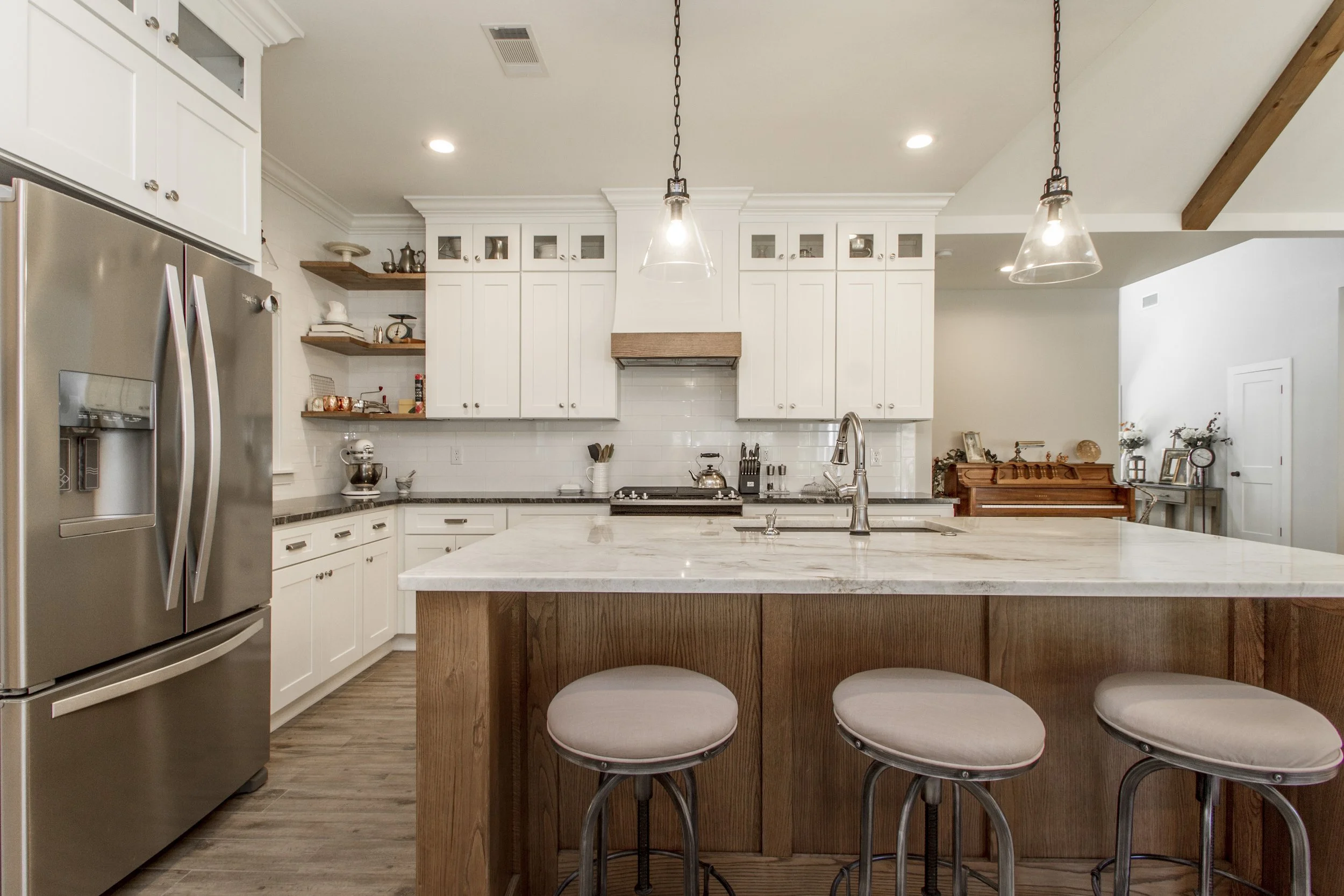 Modern kitchen with white cabinets, stainless steel refrigerator, marble countertop island with three barstools, pendant lighting, and a stove with a wooden range hood.