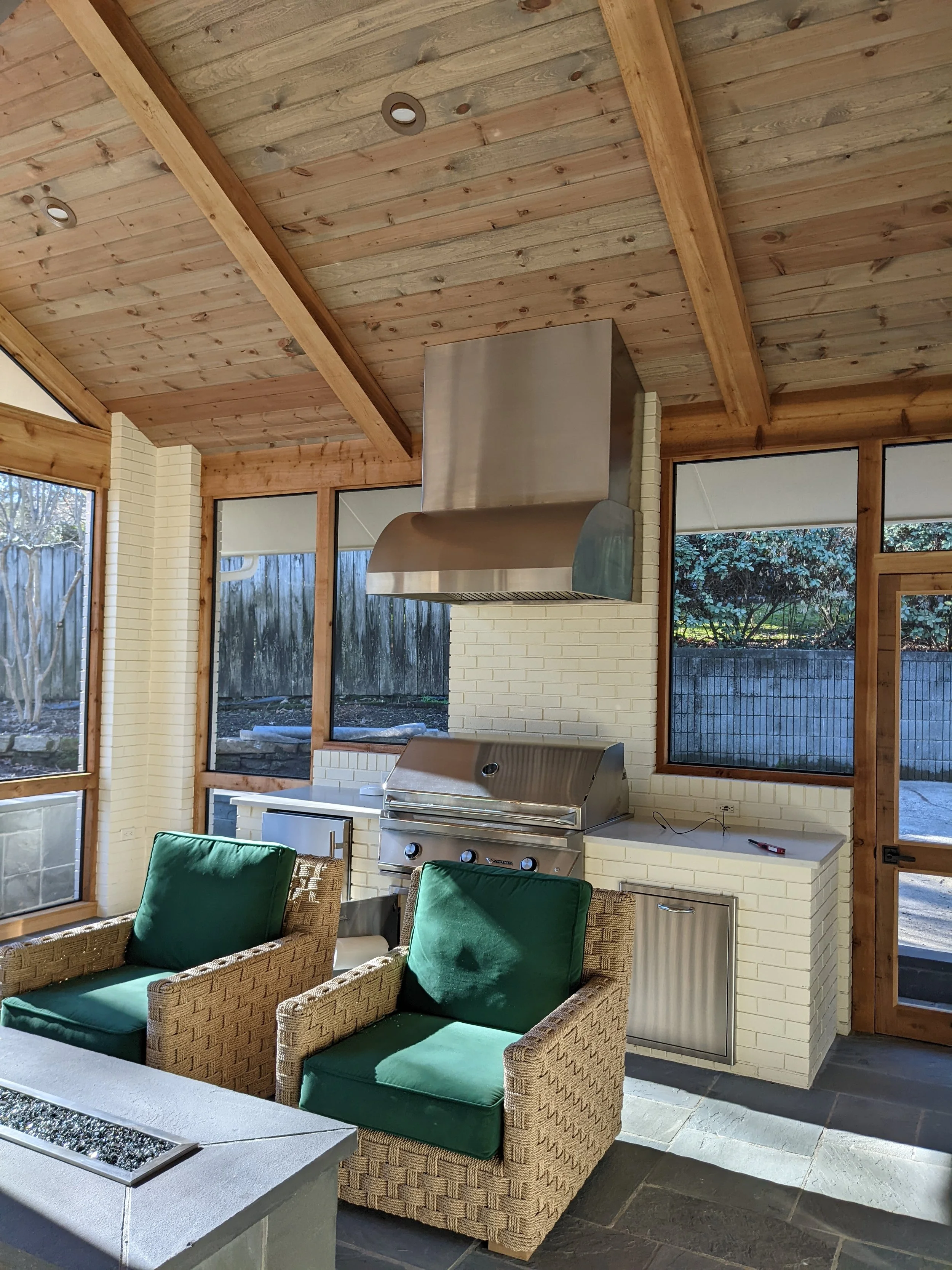 Outdoor patio area with wicker chairs with green cushions, a white brick fireplace, and a stainless steel grill beneath a wooden ceiling with large windows and sliding glass doors.