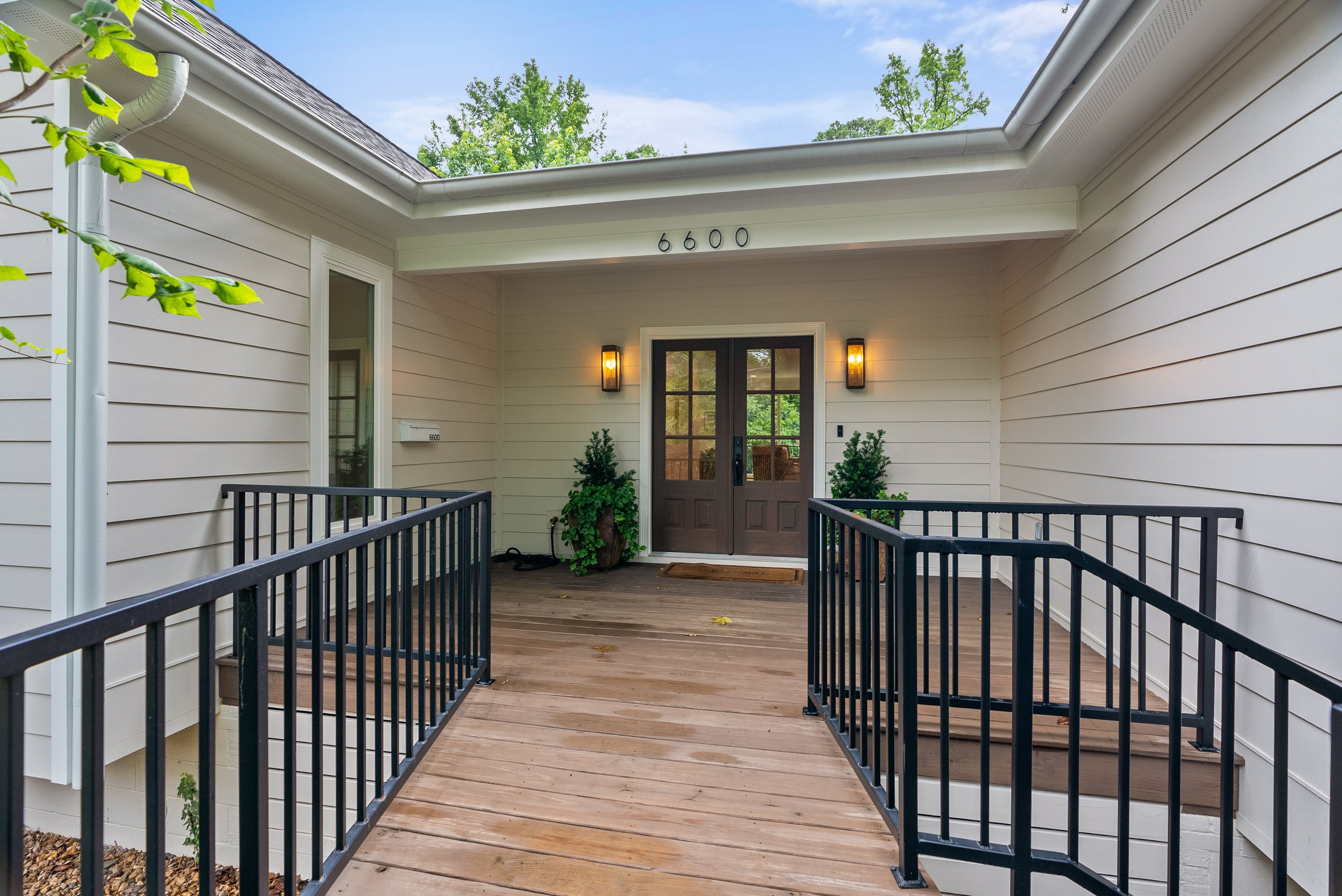 Front porch with a black railing, potted plants near a door with large glass panes, and wall-mounted lights on a beige house with horizontal siding. The house number "6600" is above the porch ceiling. The sky is partly cloudy, and trees are visible i