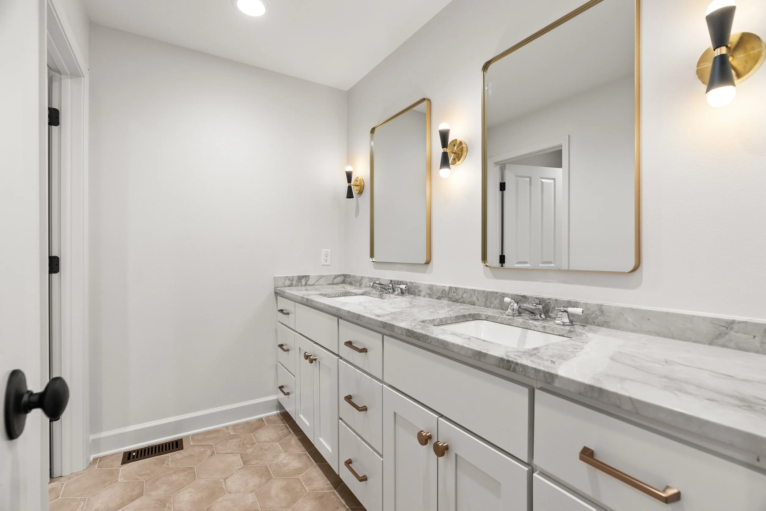 Bathroom vanity with a white cabinet, marble countertop, two sinks, and two mirrors, with gold accents on the mirrors and handles, and black sconces with gold bases.