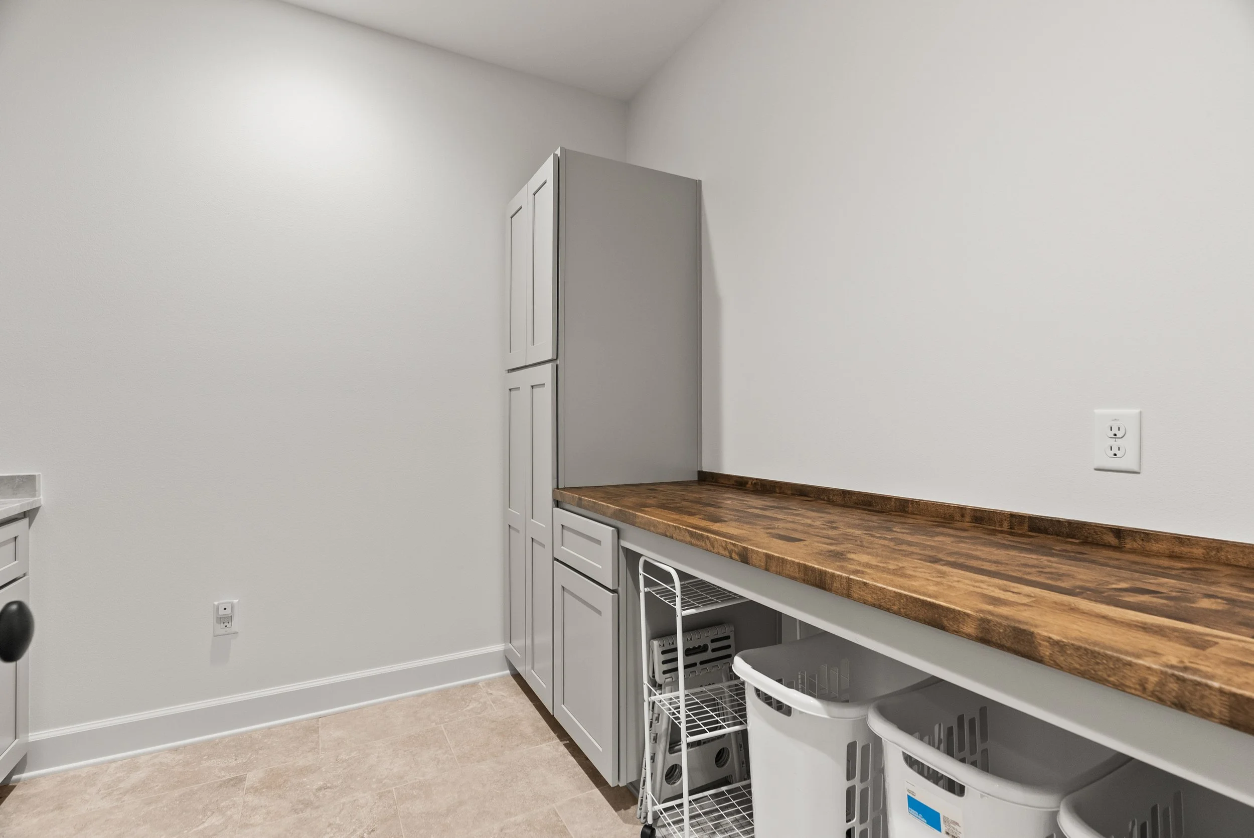A corner of a kitchen with white cabinets, wooden countertop, and a white wall with a double electrical outlet.