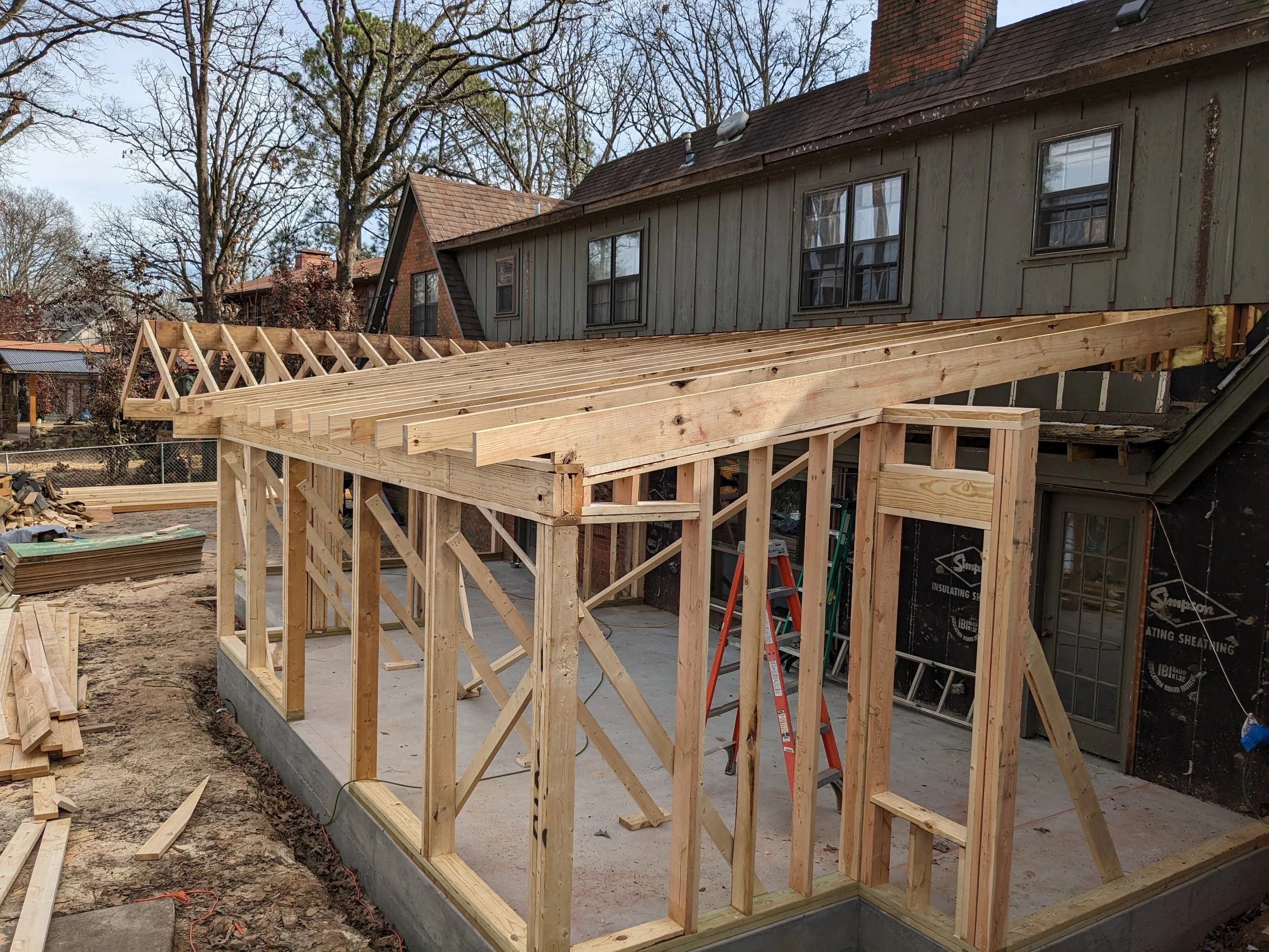 A house under construction with a wooden framework for a deck or addition in progress. There are trees in the background and construction materials on the ground.