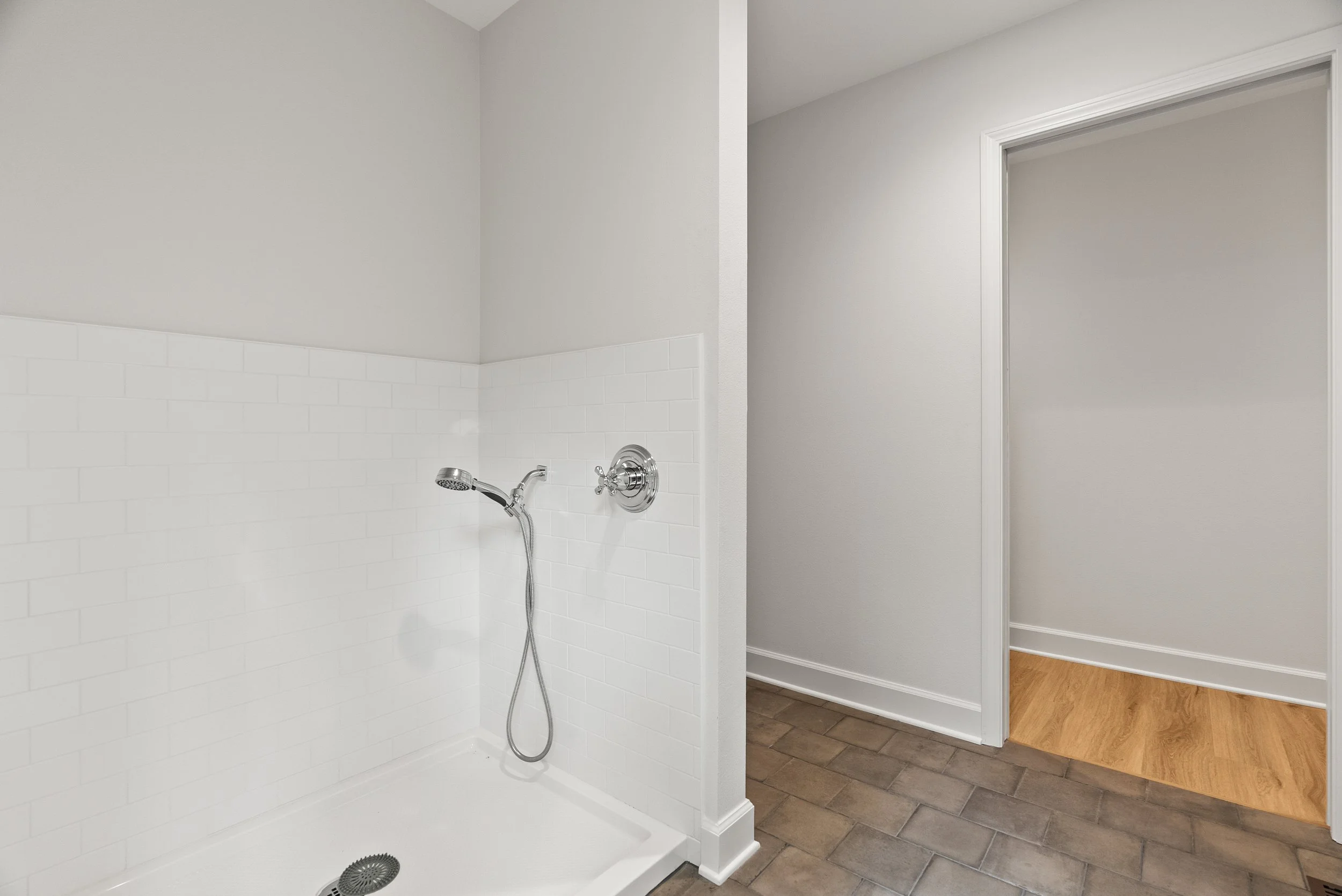 A bathroom with a white tiled shower area and a medical shower head. Adjacent to the shower is a hallway with a wood floor and a white wall.
