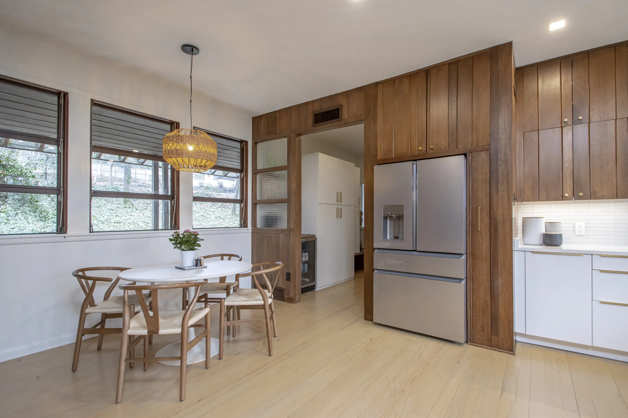 A kitchen with wooden cabinets, a refrigerator, a white countertop, and a dining area with a white round table surrounded by four wooden chairs. There is a window with blinds, a hanging wicker light, and some potted plants on the table.