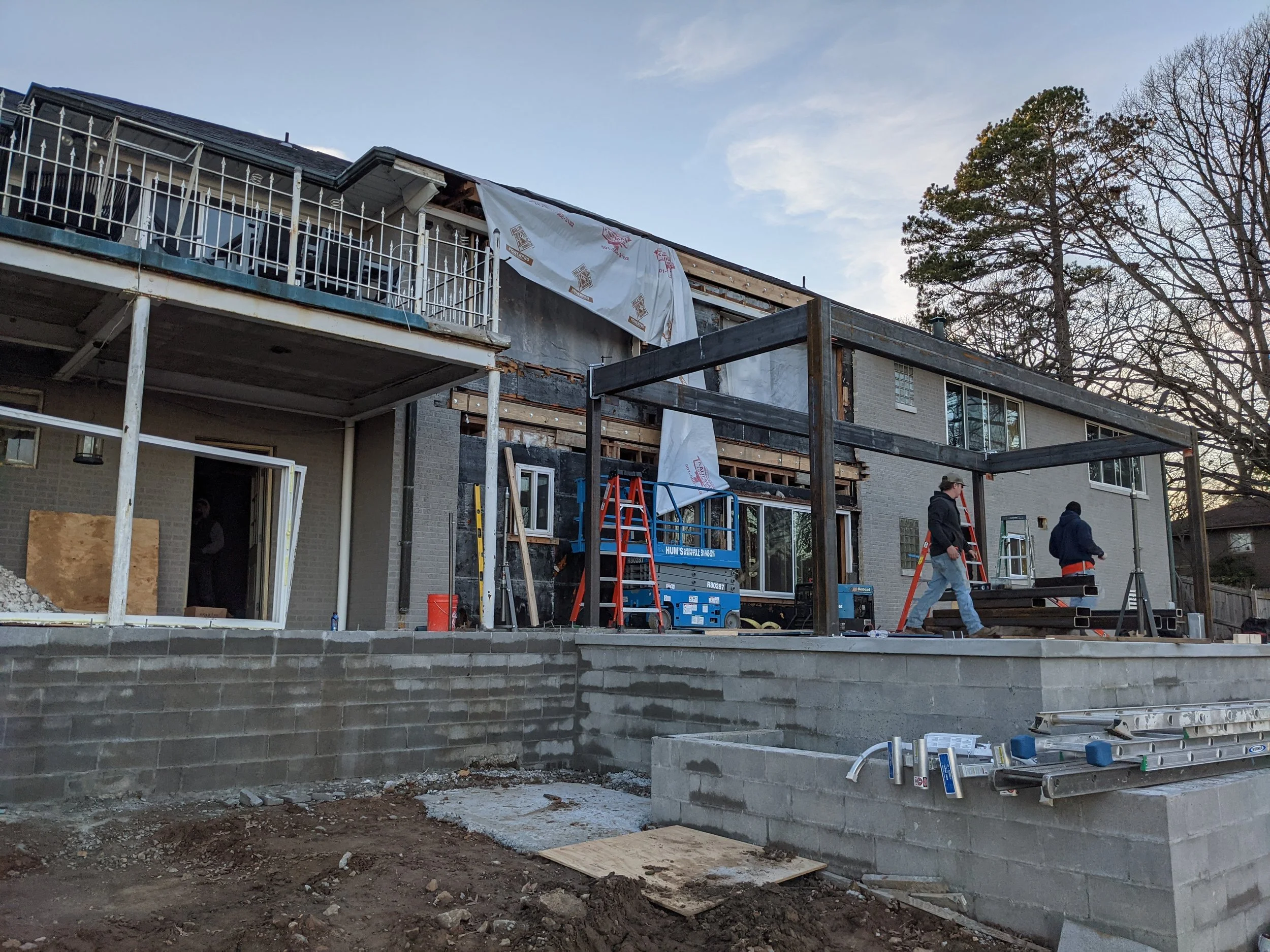 Construction workers are building a multi-story residential or commercial building outdoors. The structure's exterior walls are partially finished, with some sections showing exposed insulation and framing, and others completed with brick or siding. 
