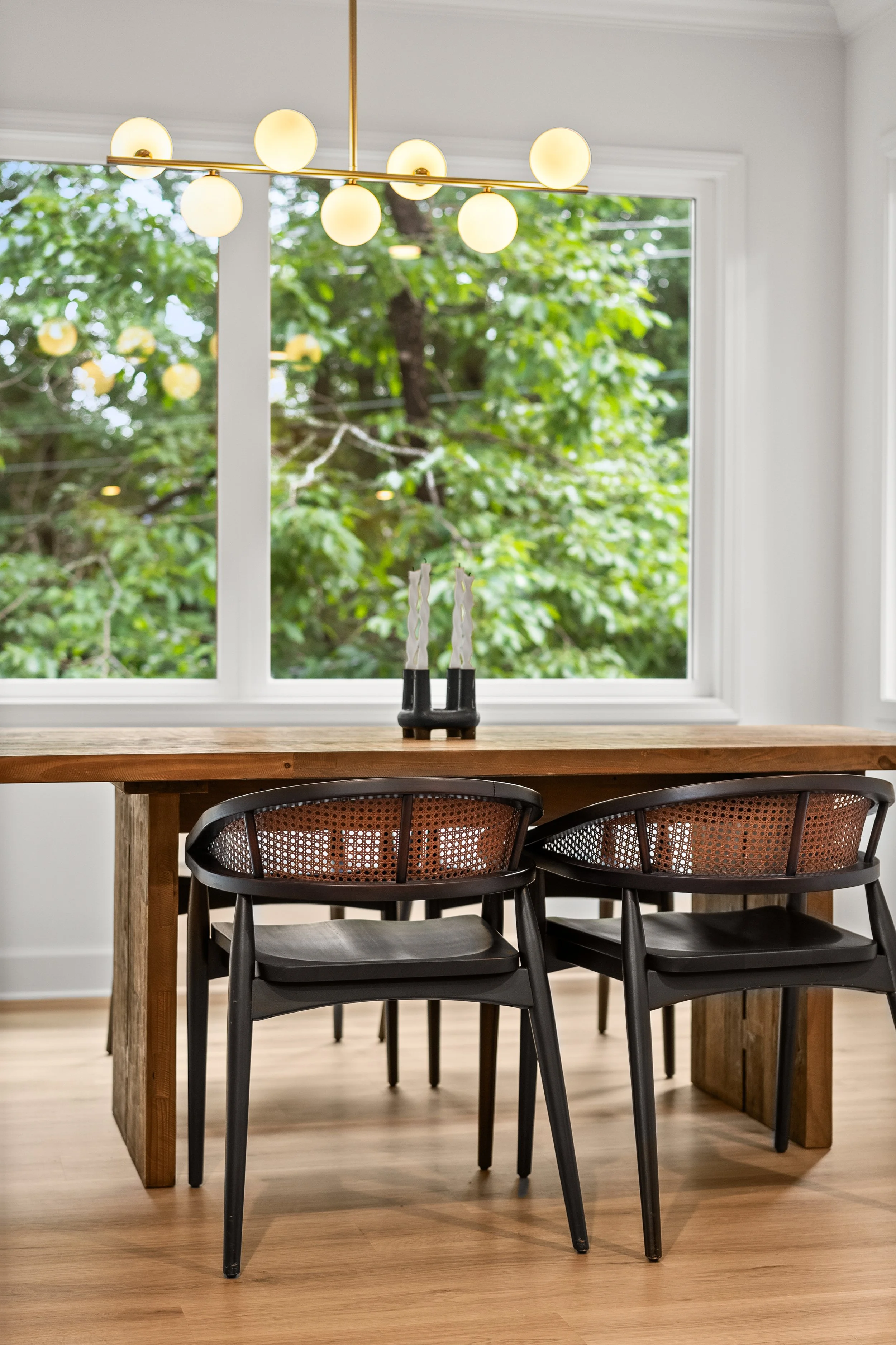 A modern dining area featuring a wooden table with black chairs, large windows showing green trees outside, and a gold chandelier with white globes hanging from the ceiling.