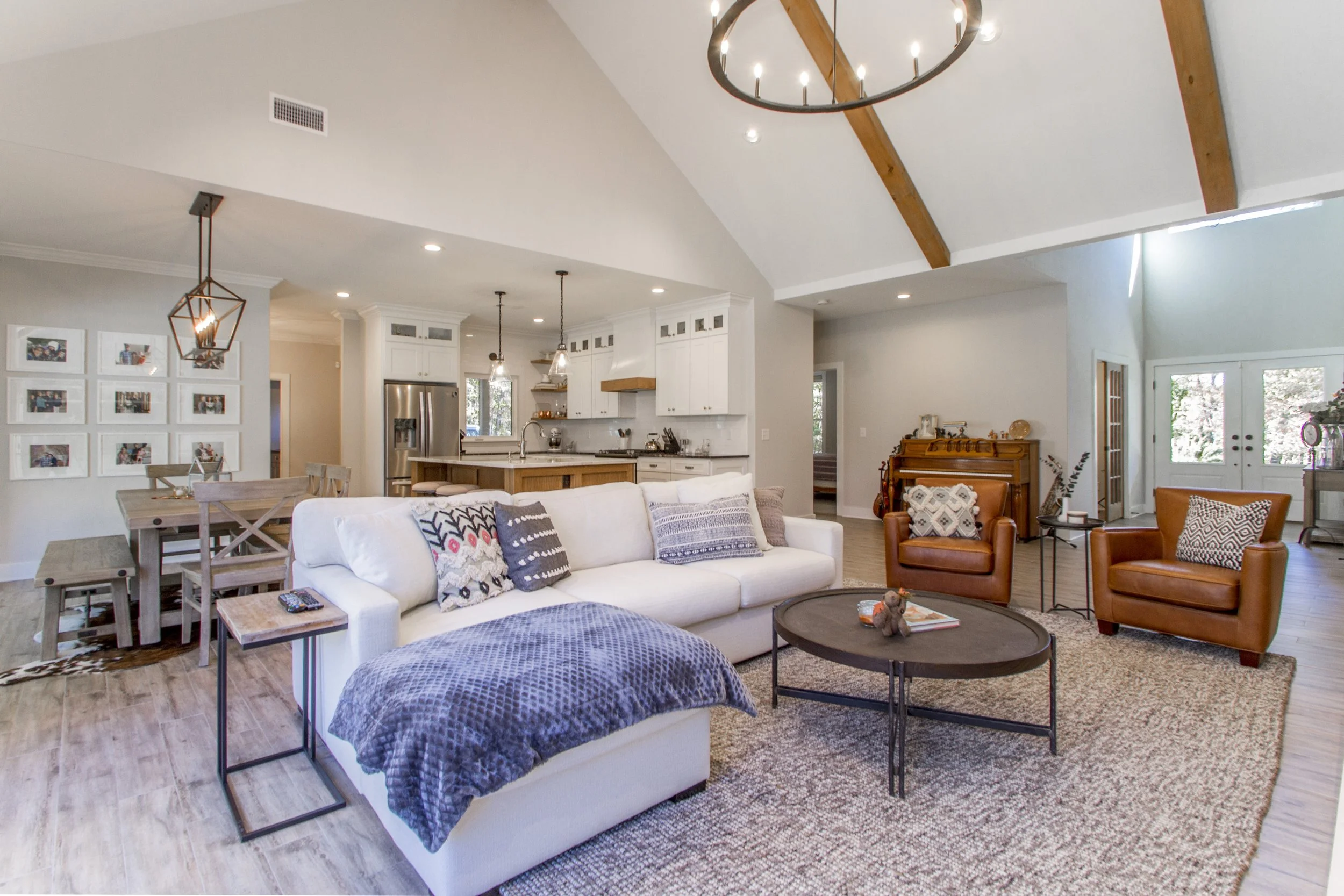Spacious open-concept living room and kitchen with vaulted ceiling, wooden beams, and large windows, decorated in a modern farmhouse style with white and neutral tones, featuring a white sofa with patterned pillows, two leather armchairs, a round cof