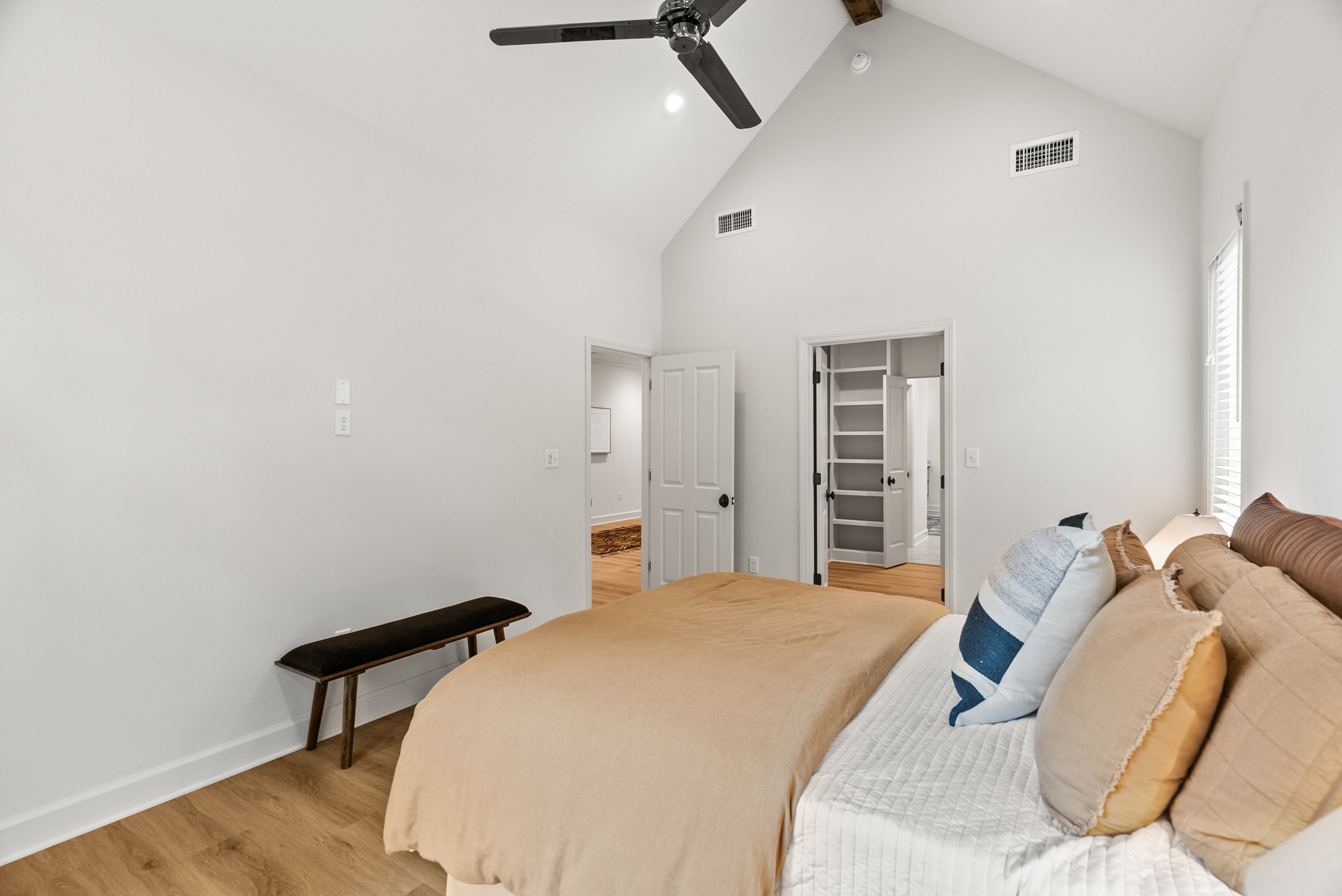 A bedroom with a white ceiling, a black ceiling fan, and white walls. There are two air vents on the ceiling, an open door leading to another room, and a window with blinds. A bed with a beige blanket and pillows is partially visible, along with a lo
