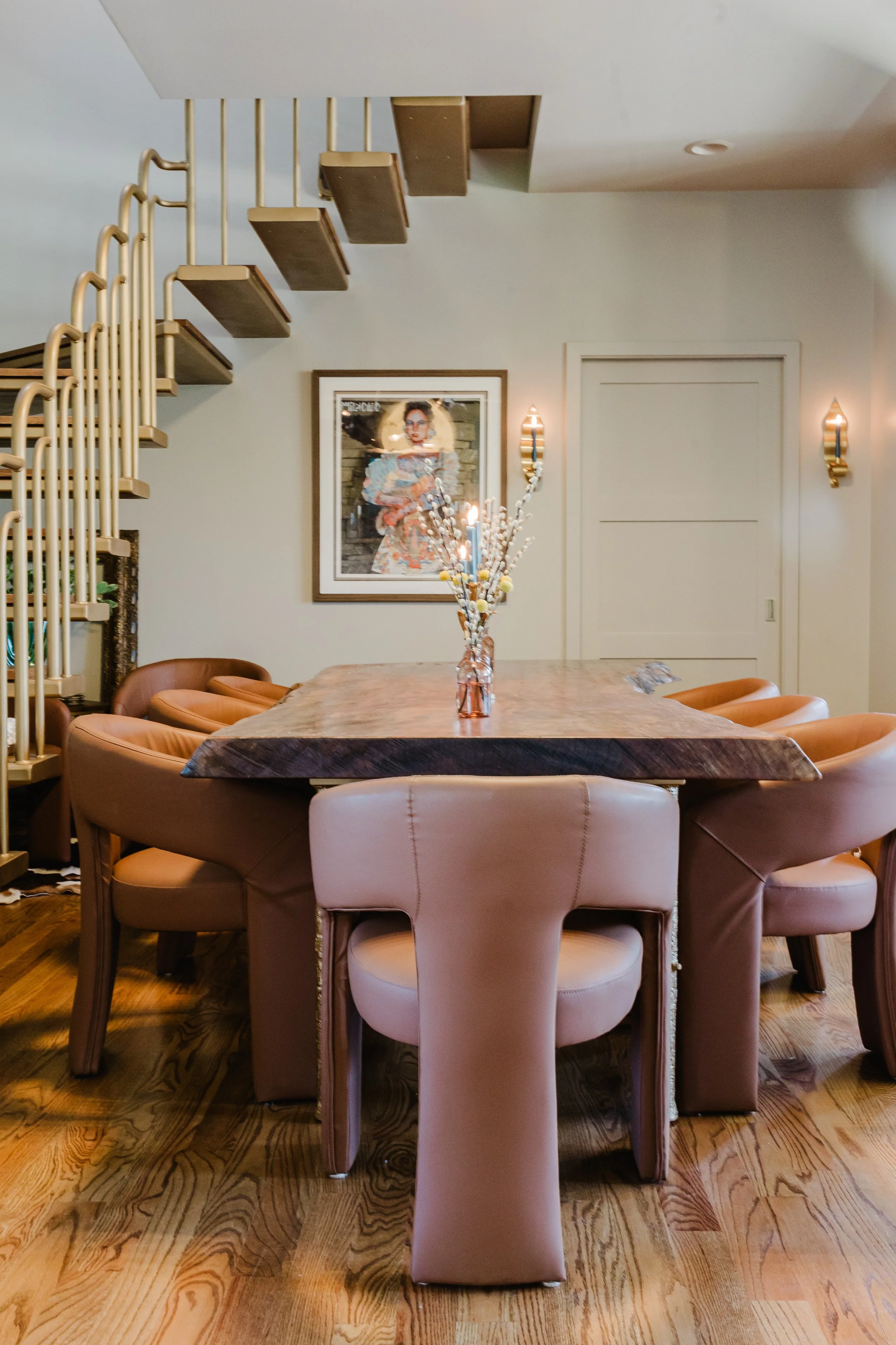 A dining room with a large wooden table surrounded by nine pink upholstered chairs. In the center of the table is a vase with decorative branches. A framed portrait of a woman hangs on the wall, with two wall-mounted lights on either side of the port
