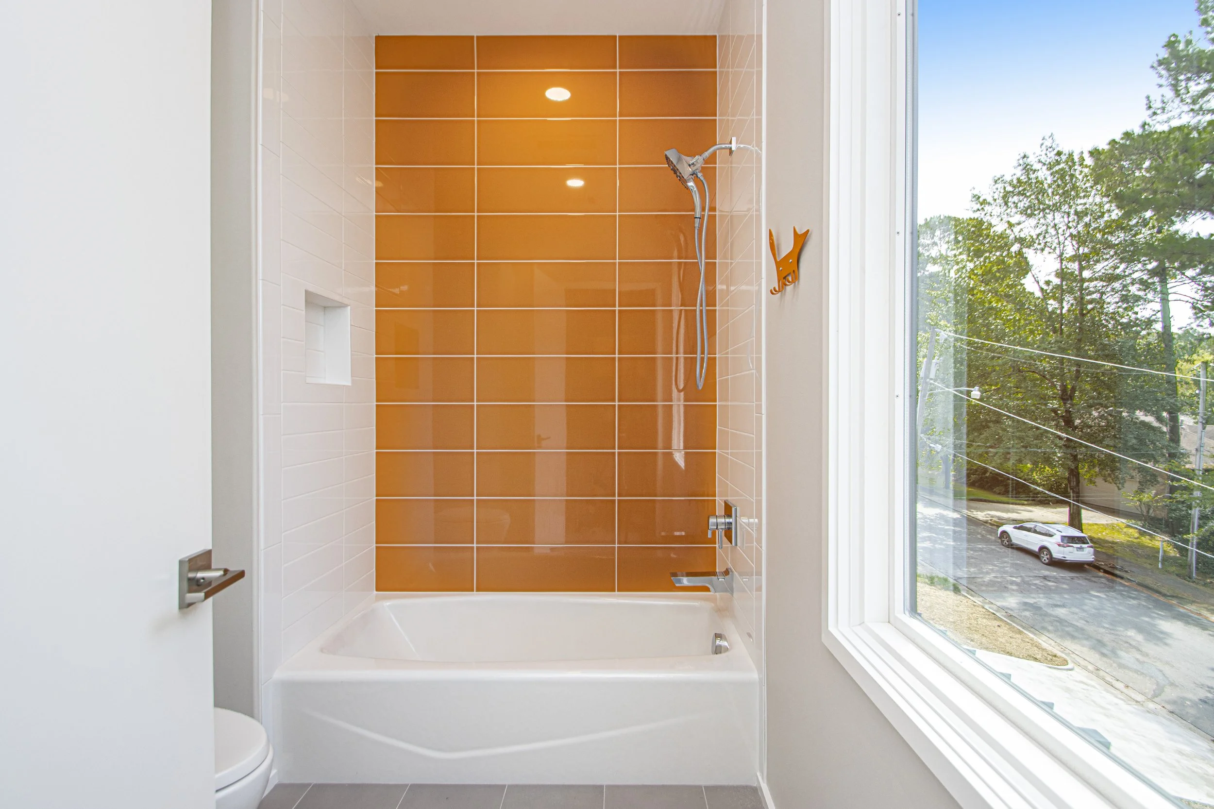 A bathroom with a shower area that has orange tiles on the back wall and white tiles on the side walls, a large window with a view of trees and a parking lot, a showerhead, and a small built-in shelf in the wall.