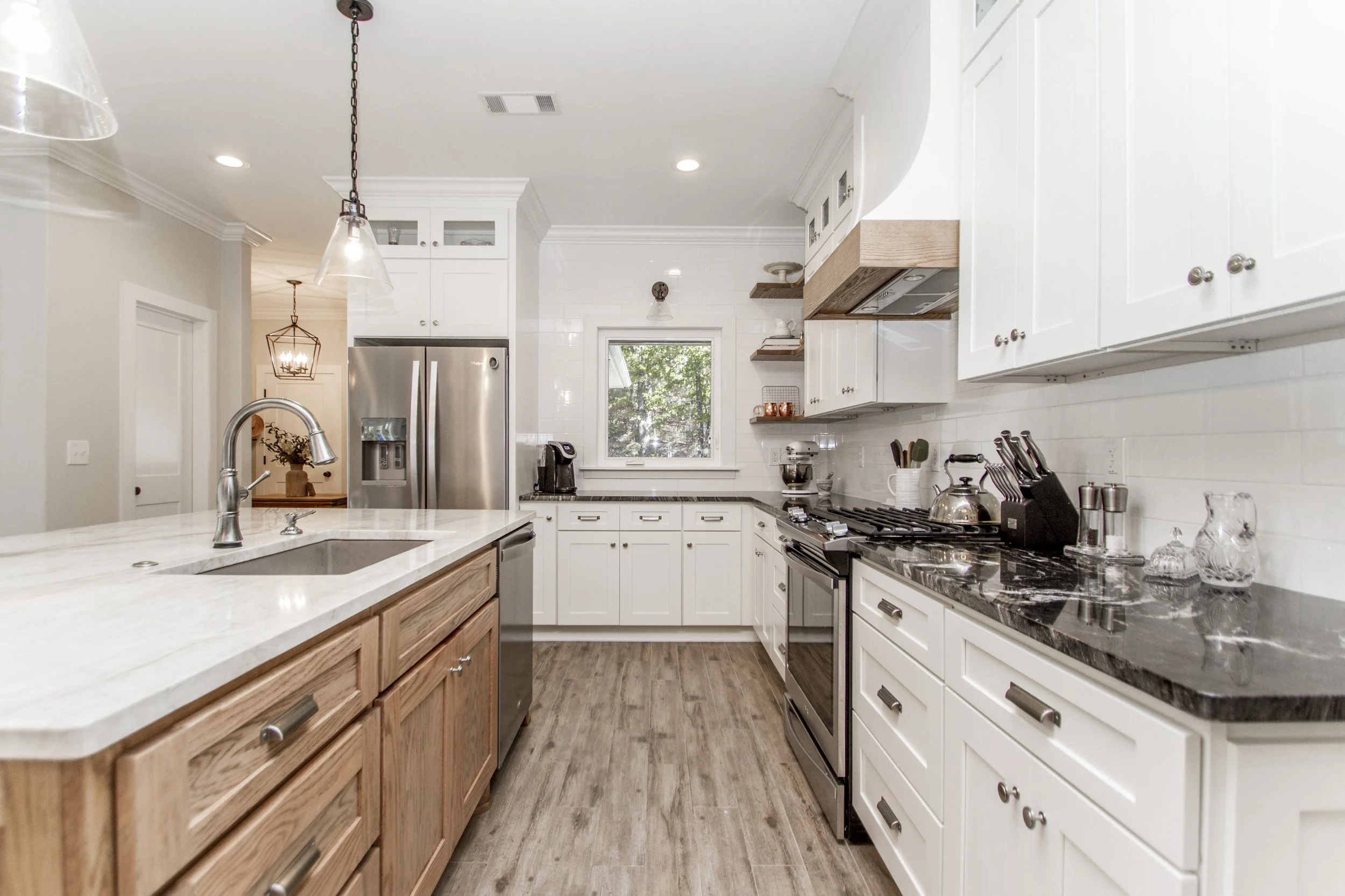 A modern kitchen with white cabinets, stainless steel appliances, black granite countertops, and a mix of wood and white finishes. There is a window overlooking trees, pendant lighting, and various kitchen utensils and appliances on the counter.