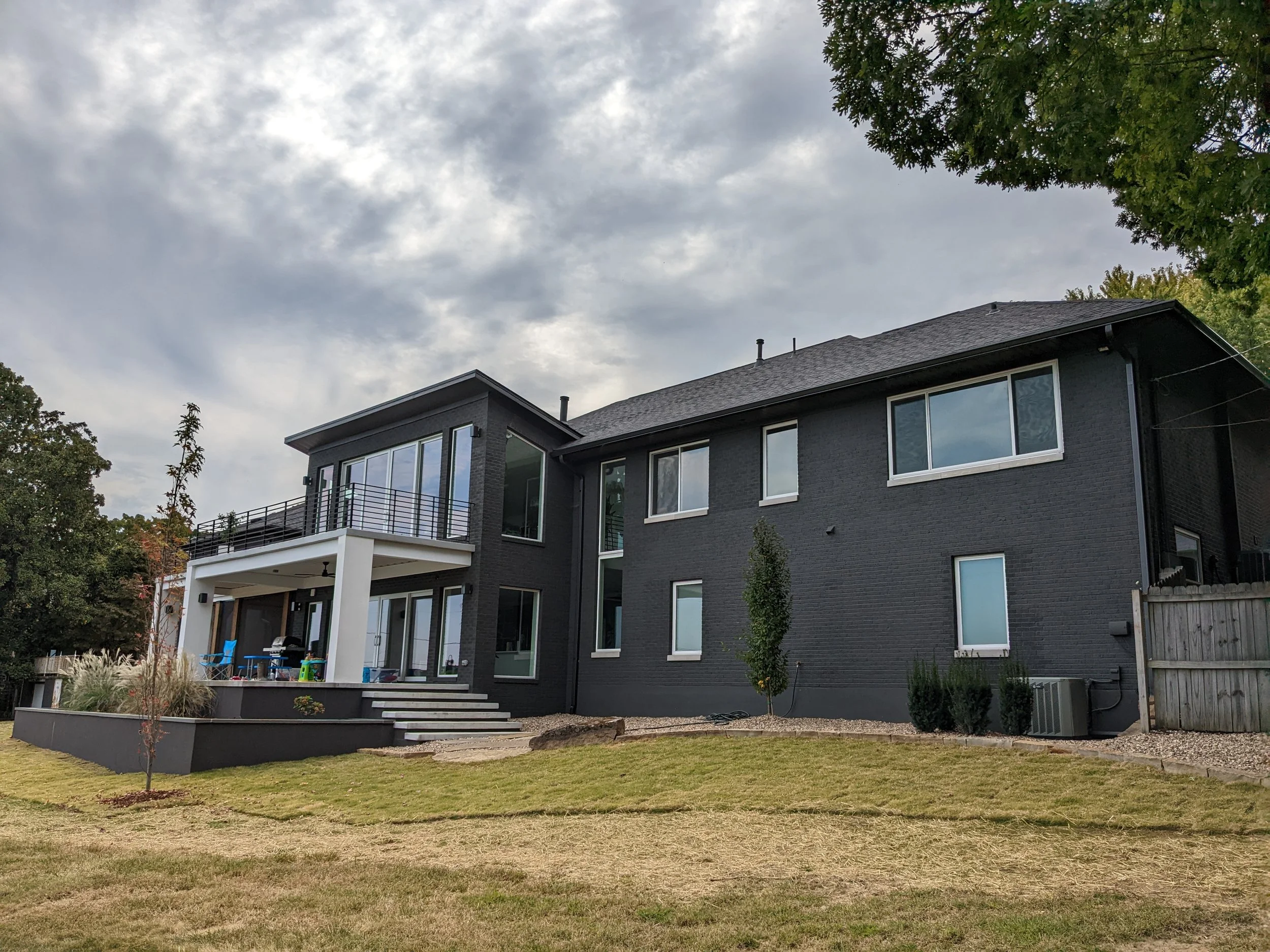 A modern two-story house with large windows, a balcony, and outdoor furniture, surrounded by a landscaped yard with grass, small trees, and plants, under a cloudy sky.