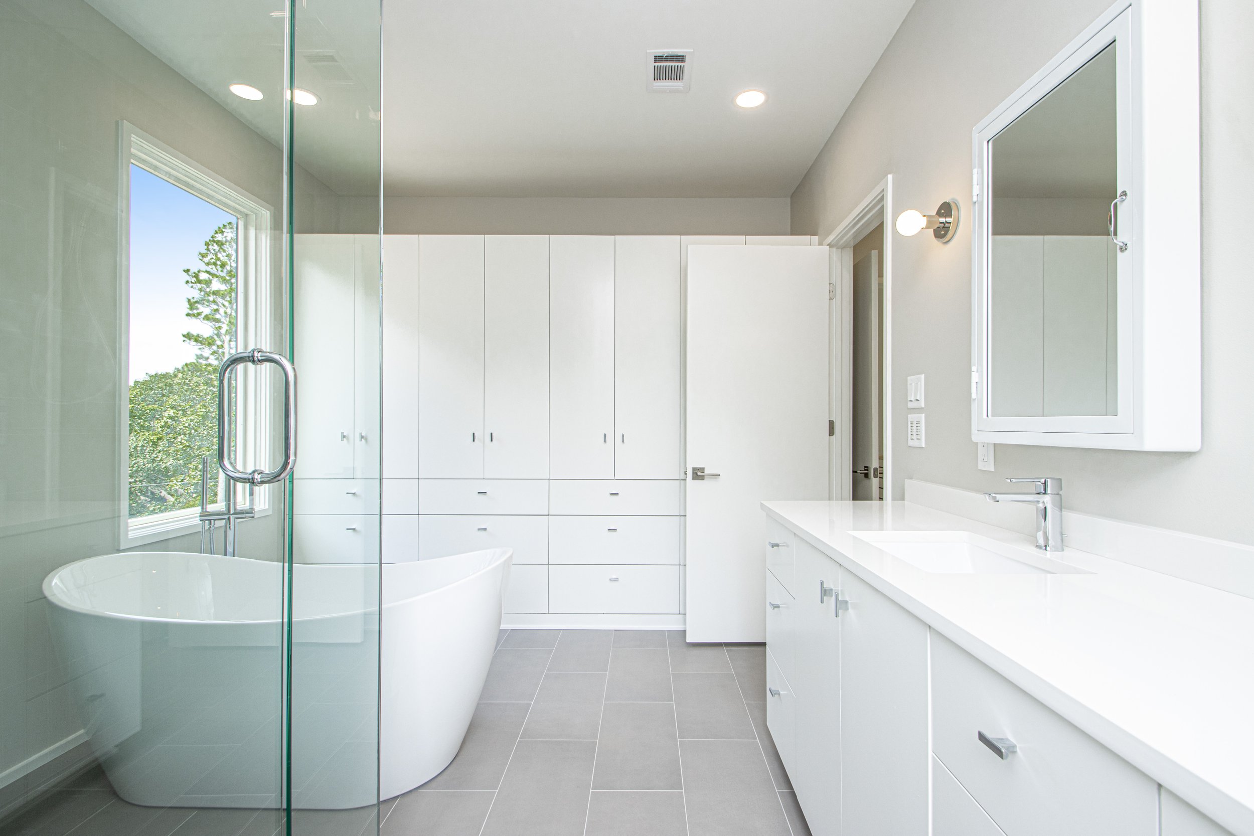 Modern bathroom with a walk-in glass shower, freestanding bathtub, white cabinetry, gray floor tiles, and a window showing trees outside.