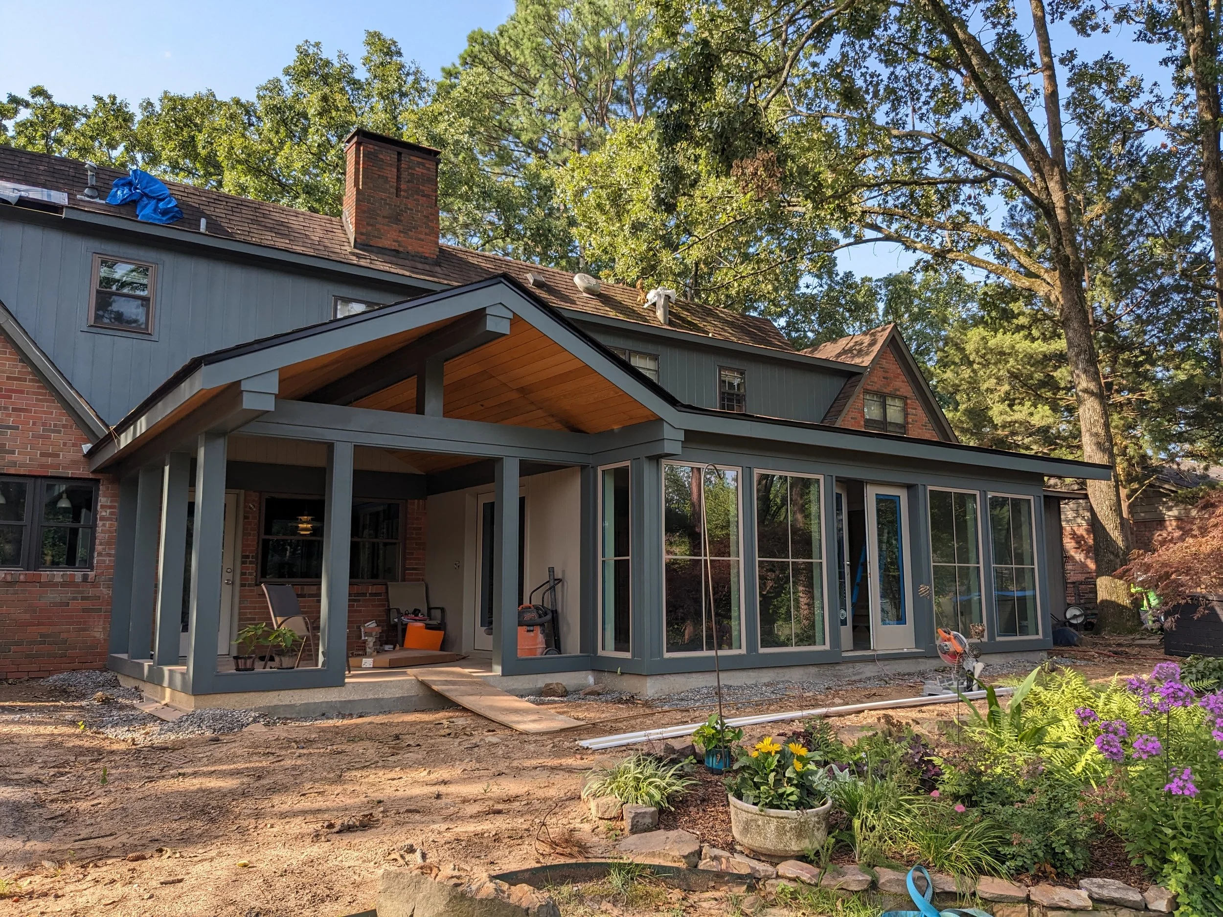 A house under construction with a screened-in porch and a sloped roof. The backyard features a flower bed with colorful flowers and a small garden area. Tall trees surround the house, and building materials are visible on the ground.