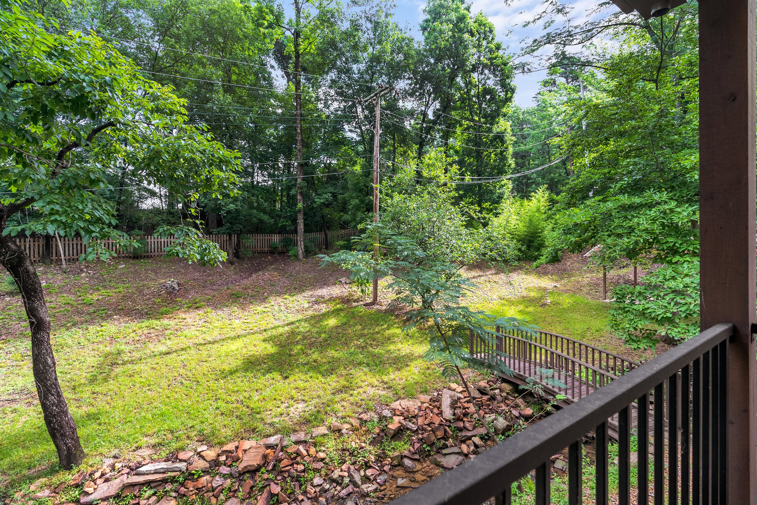 View of a backyard with green grass, trees, and wooden fence, seen from a porch with black railing and a wooden post.