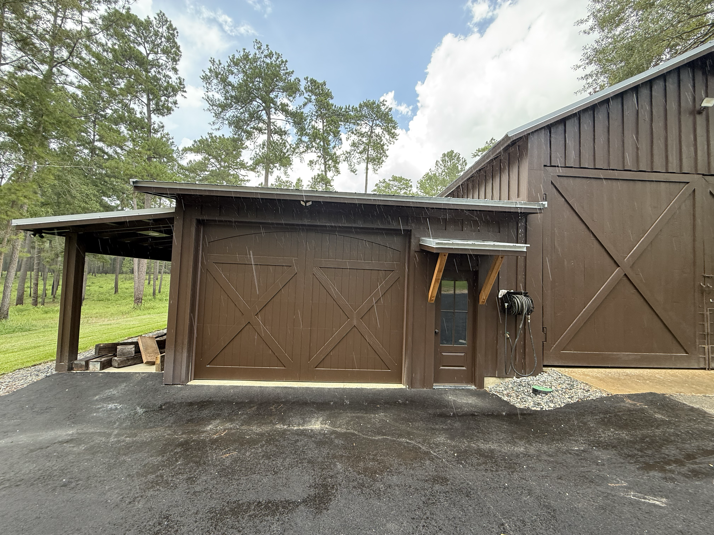 A brown wooden barn with large double doors, part of a side extension, and a hose reel attached on the right side. The barn is in a grassy area with trees in the background, and it's raining with visible raindrops falling.