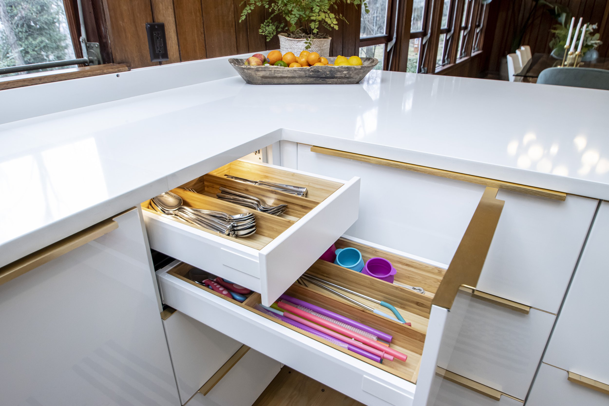 Open kitchen drawer with silverware, kitchen tools, and children's toys inside, with a white countertop and wooden accents.