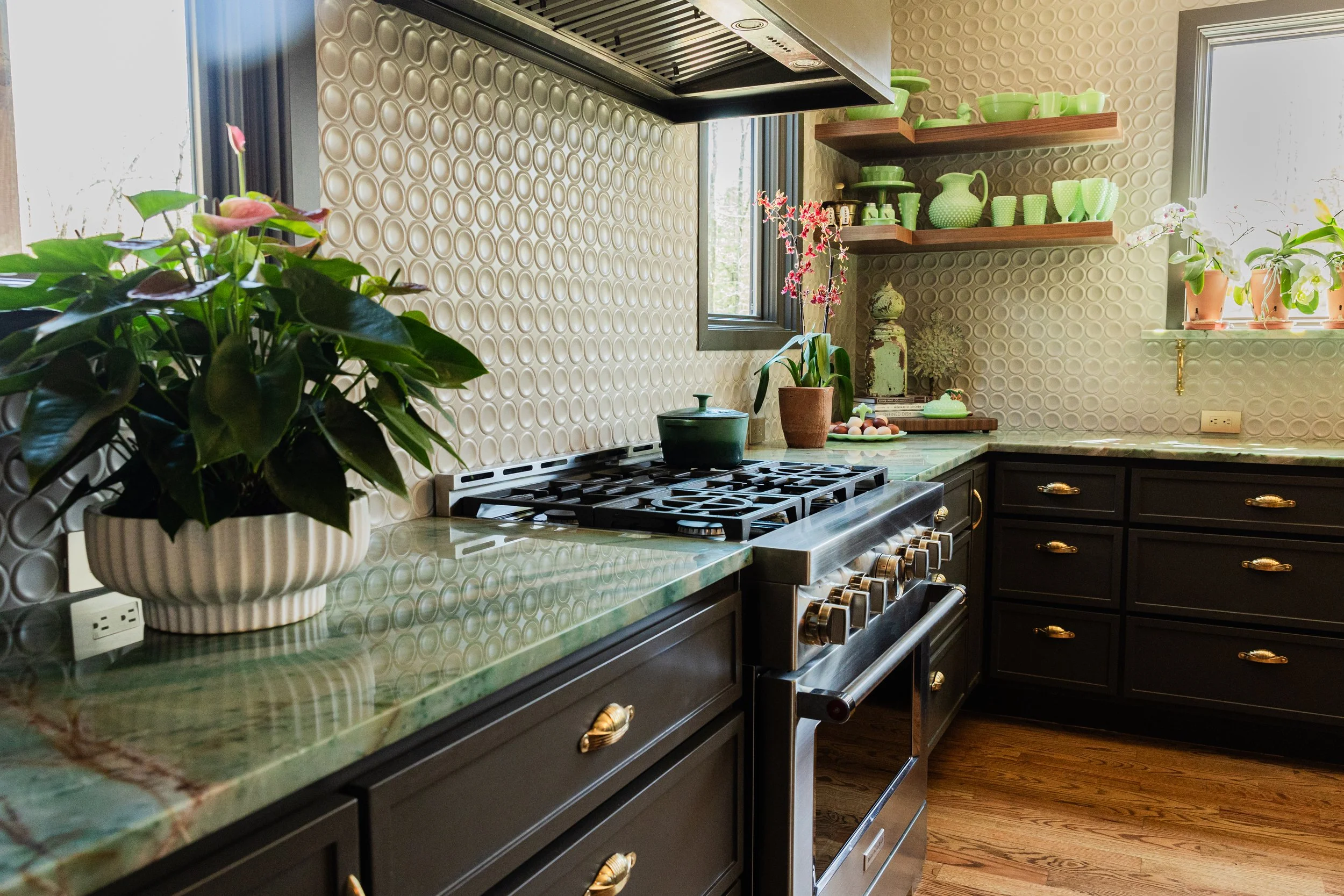A kitchen with a green marble countertop, dark cabinets with gold handles, a stove, and two shelves filled with green dishware and pottery. There are two potted plants on the windowsill and a pink flowering plant on the counter. Light is coming throu