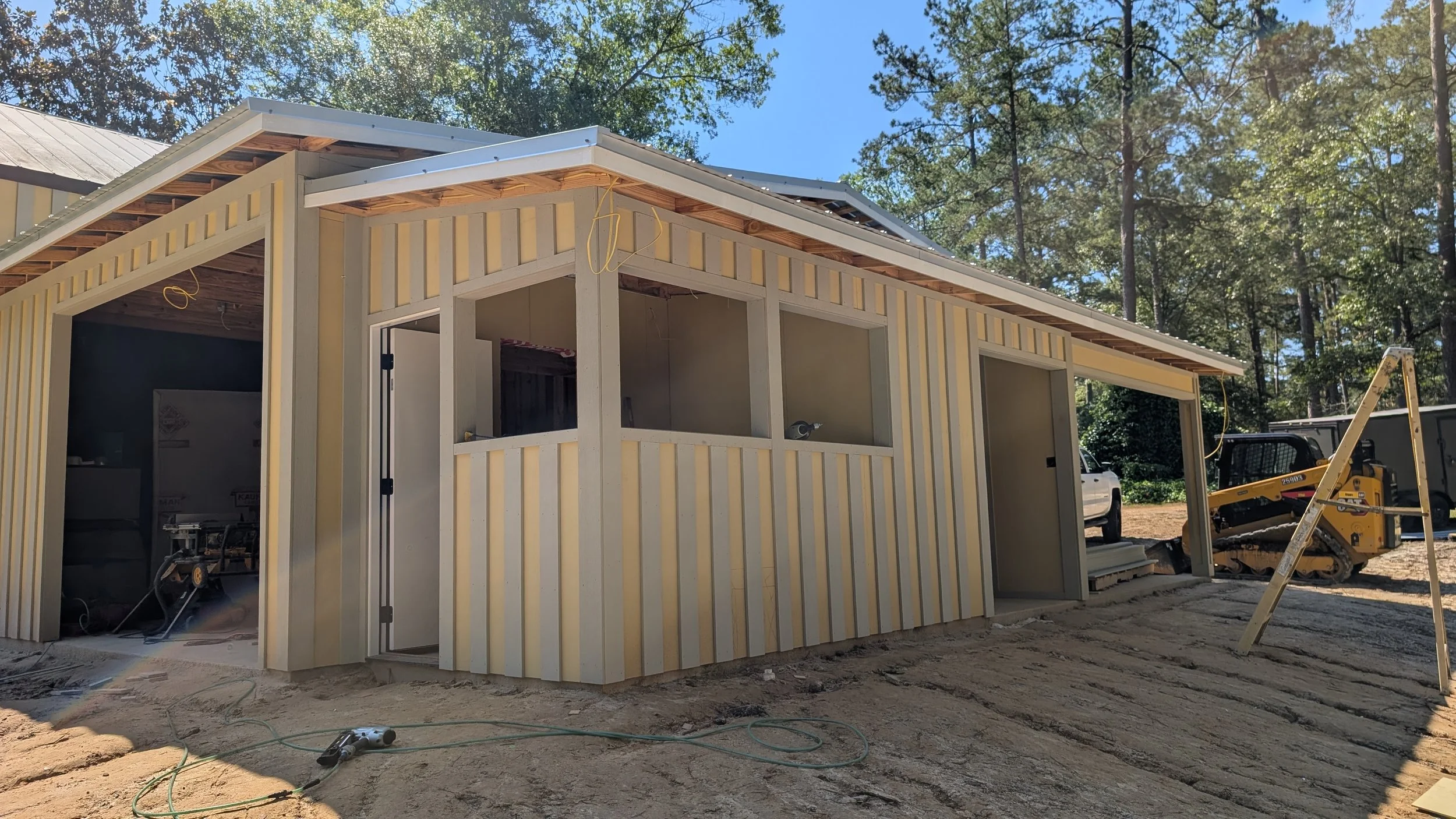 Under construction, single-story building with yellow vertical siding, open windows, construction tools, and machinery, surrounded by trees.