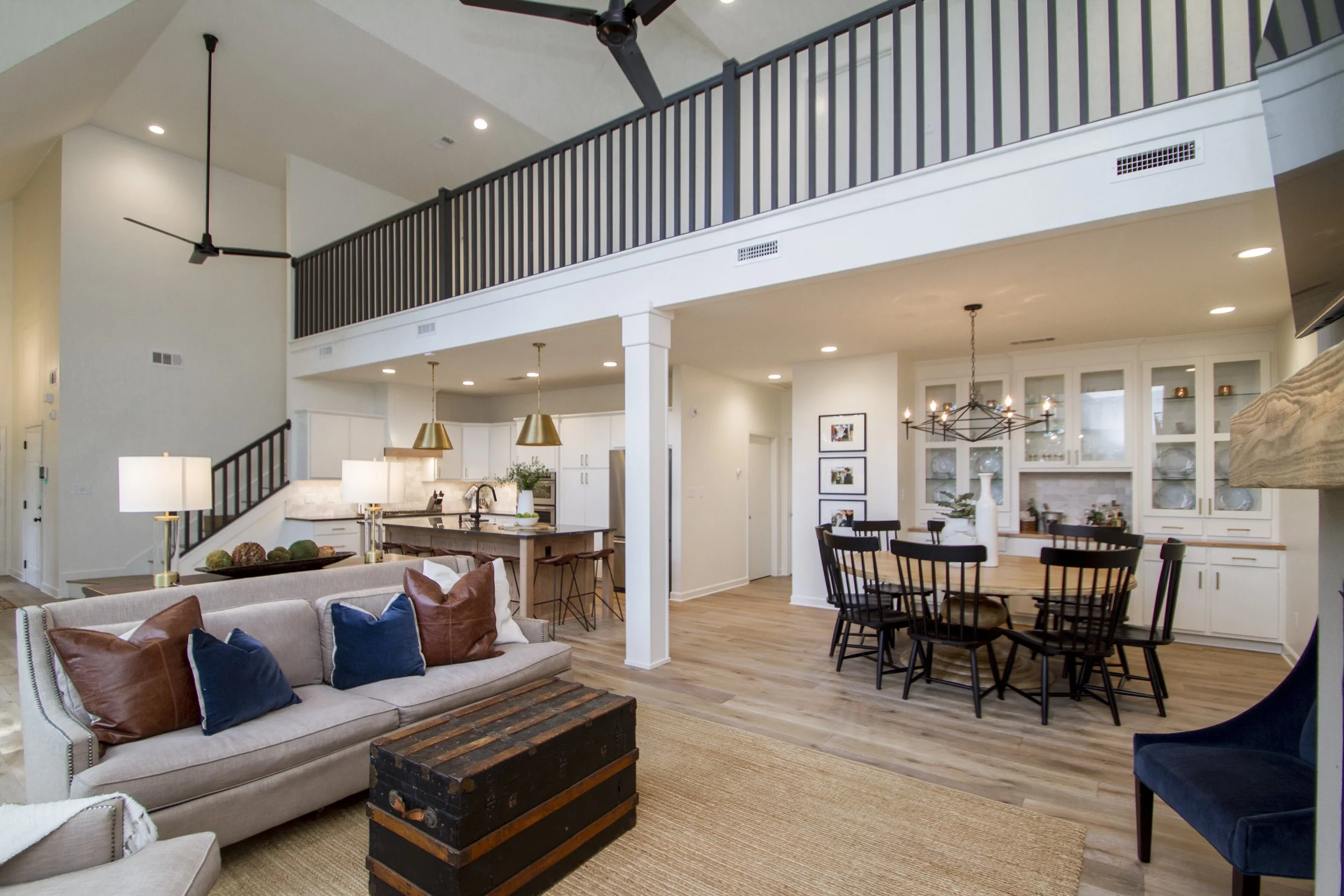 Open-concept living space with a beige sofa, blue and brown pillows, a rustic coffee table, a dining area with black chairs, white cabinetry, and high ceilings with modern lighting and ceiling fans.