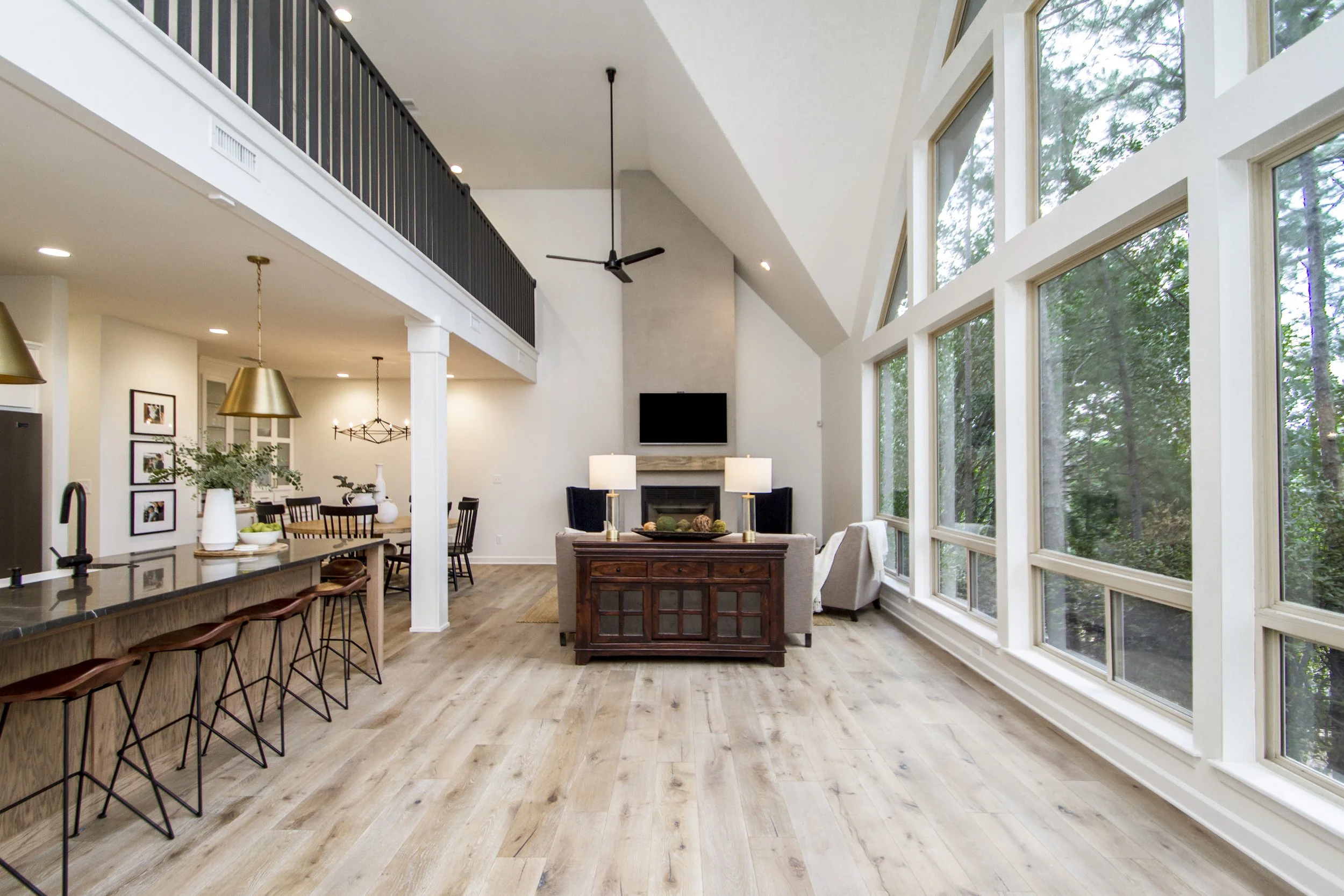 Open-concept living room and kitchen area with large windows showing trees outside, wood floors, and modern furniture.