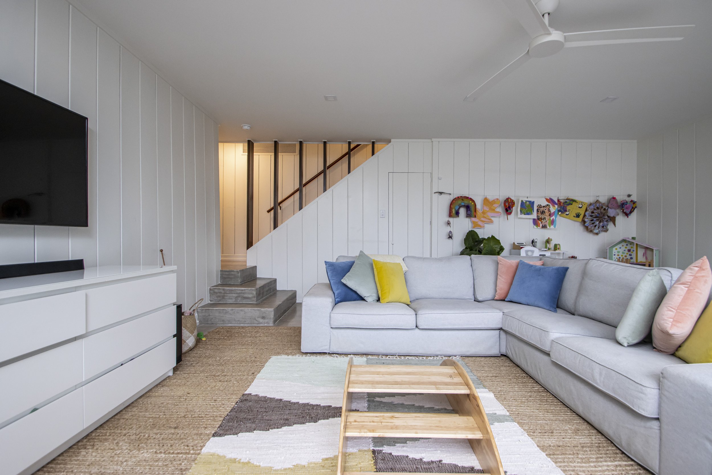 Living room with white walls, a white sofa with colorful pillows, a wall decorated with children's artwork, a TV on the left, and stairs with dark wood railings in the background.