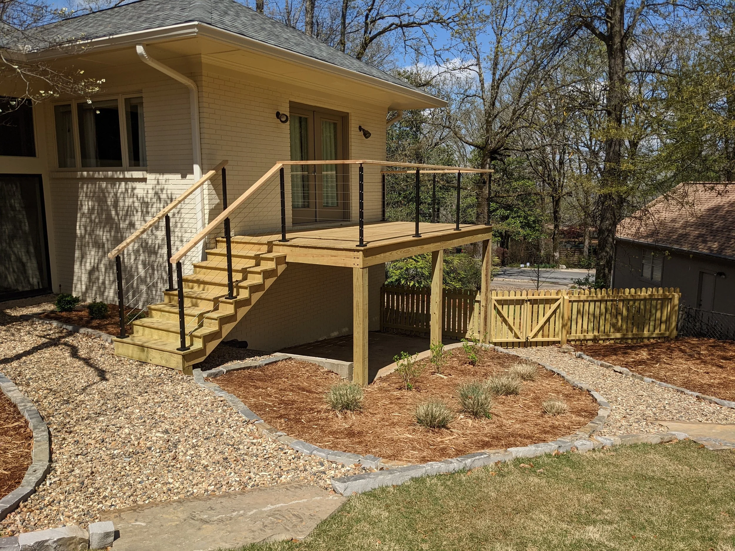 Backyard with a raised wooden deck, black metal railing, sliding glass door, and exterior wall of a house, surrounded by trees and landscaped garden beds with mulch and small plants.