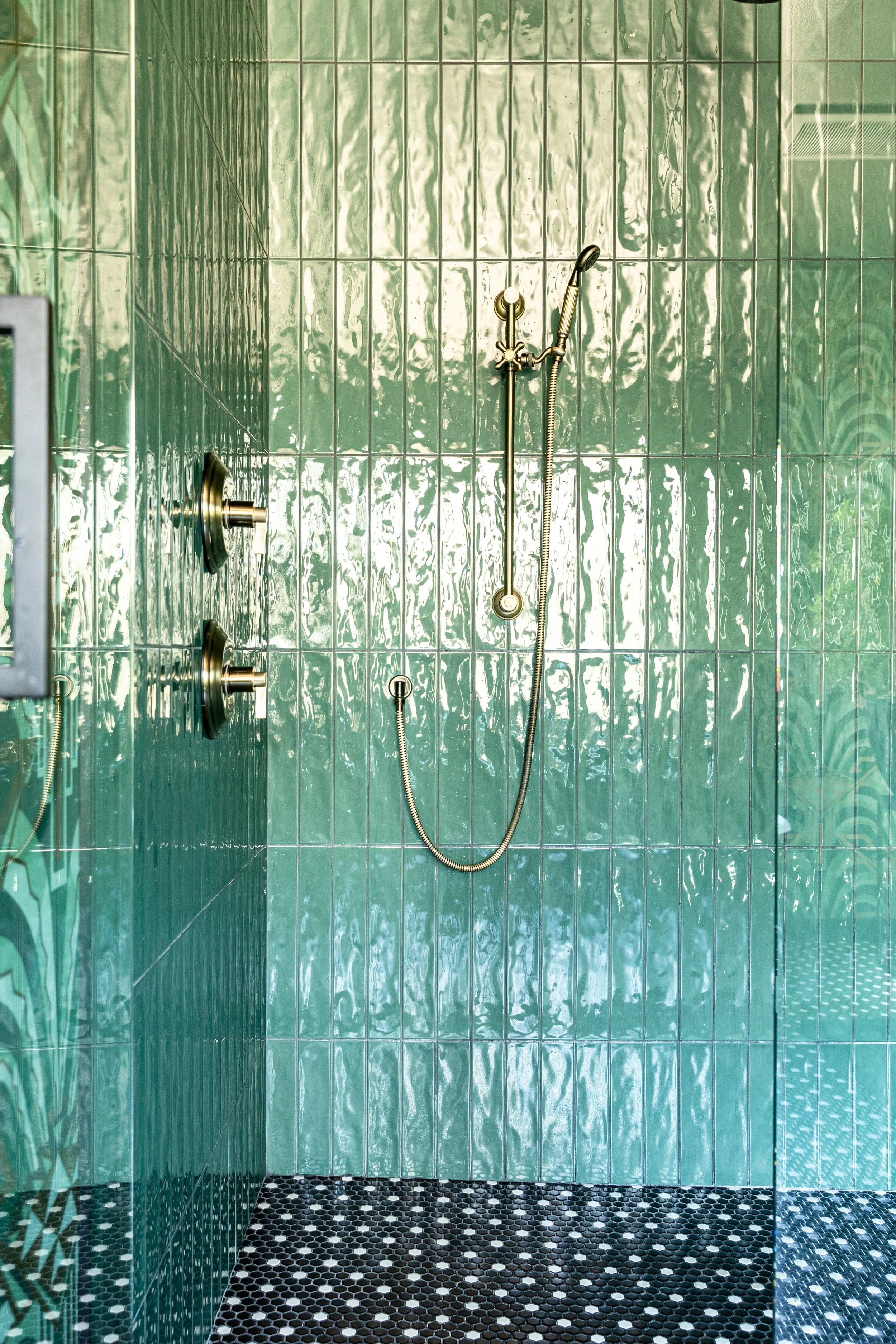 Green-tiled shower with black and white hexagonal floor, brass shower fixtures, handheld showerhead, and glass door.