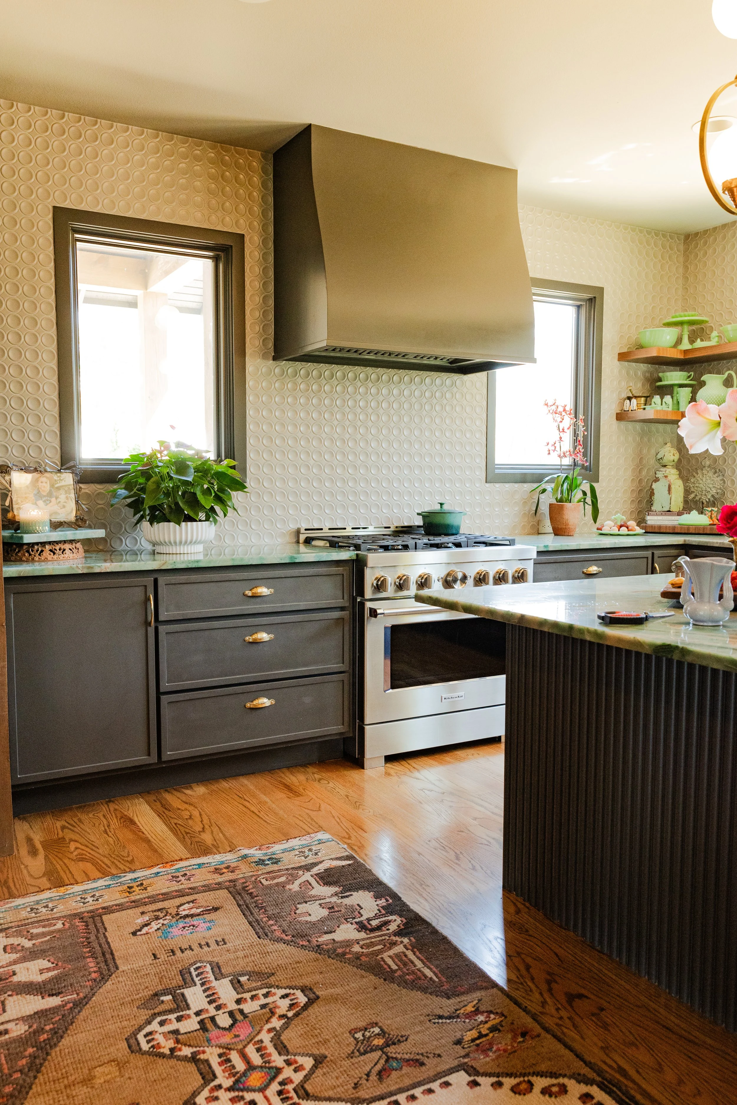 A cozy kitchen with dark gray cabinets, a white gas stove, and a beige textured backsplash. Two windows with natural light, plants on the counter, and open shelves with green dishes and decorative items. A patterned rug on wooden flooring.