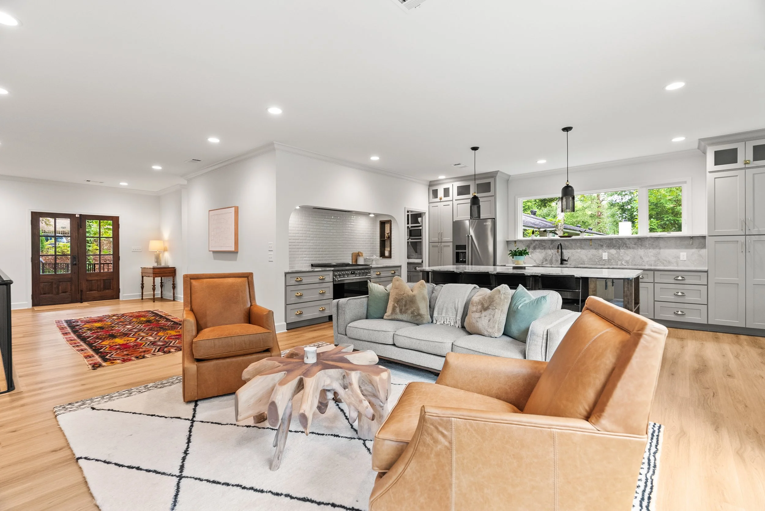 Open-concept living room and kitchen with white walls, hardwood floors, leather armchairs, a gray sofa with pillows, a wooden coffee table, a colorful rug, kitchen cabinets, stainless steel refrigerator, and large windows showing greenery outside.
