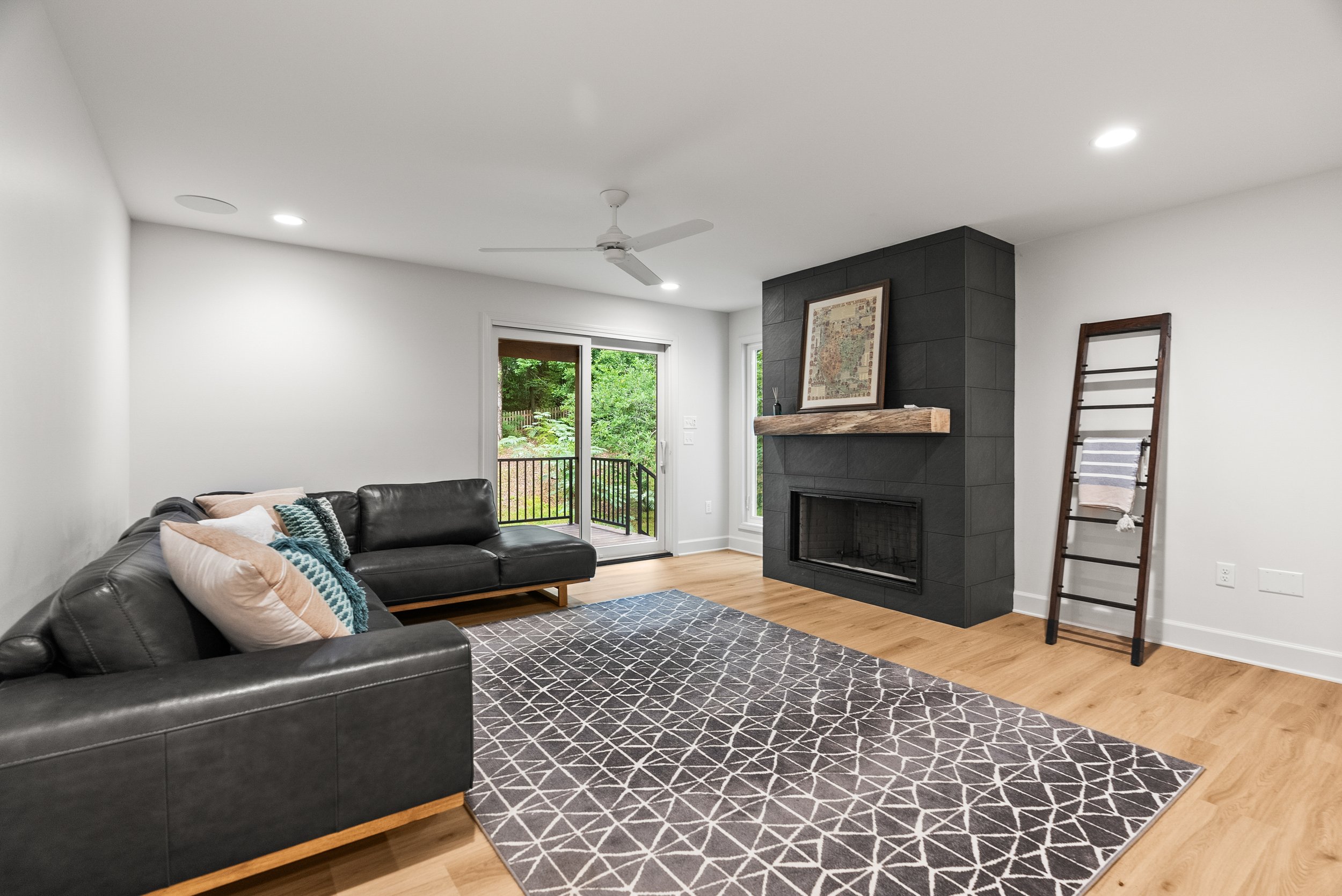 A living room with white walls, a black leather sofa with pillows, a patterned rug, a modern black fireplace with a wooden mantel, a framed artwork above the fireplace, a wooden ladder decor, and large sliding glass doors opening to a green outdoor s