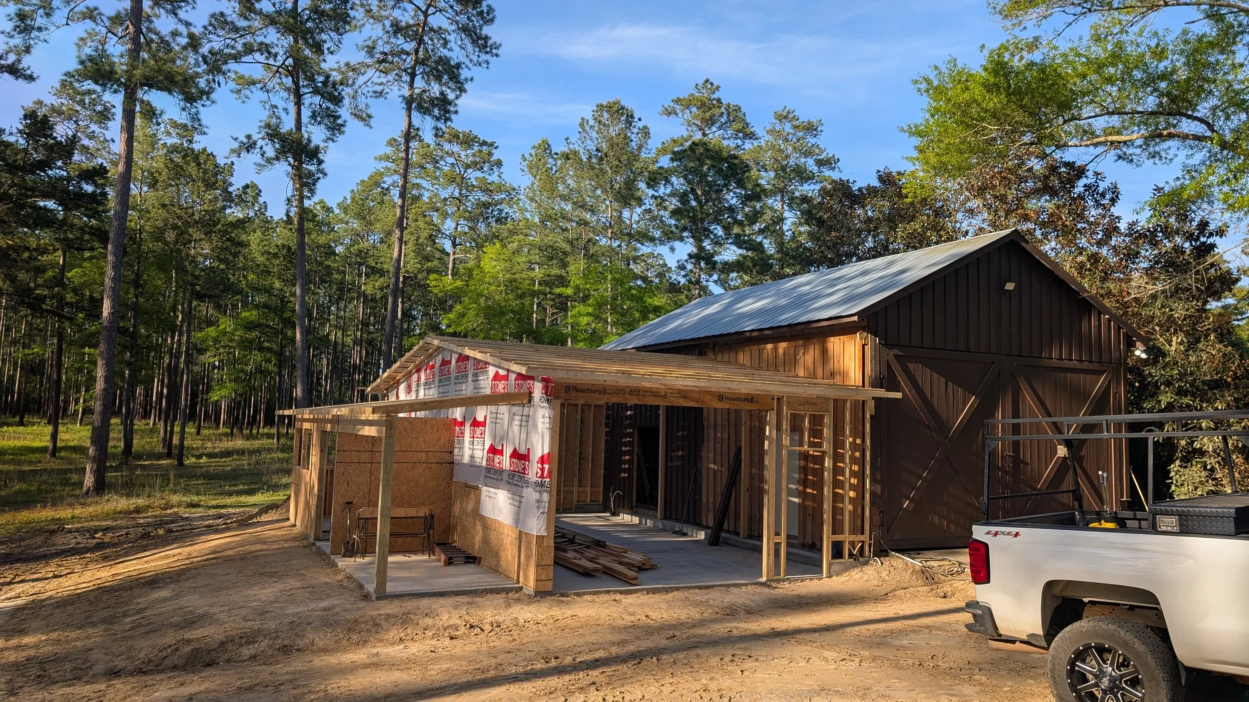 A barn under construction with a wooden frame and partially covered with weather-resistant sheathing, situated in a wooded area with tall trees and clear blue sky in the background.