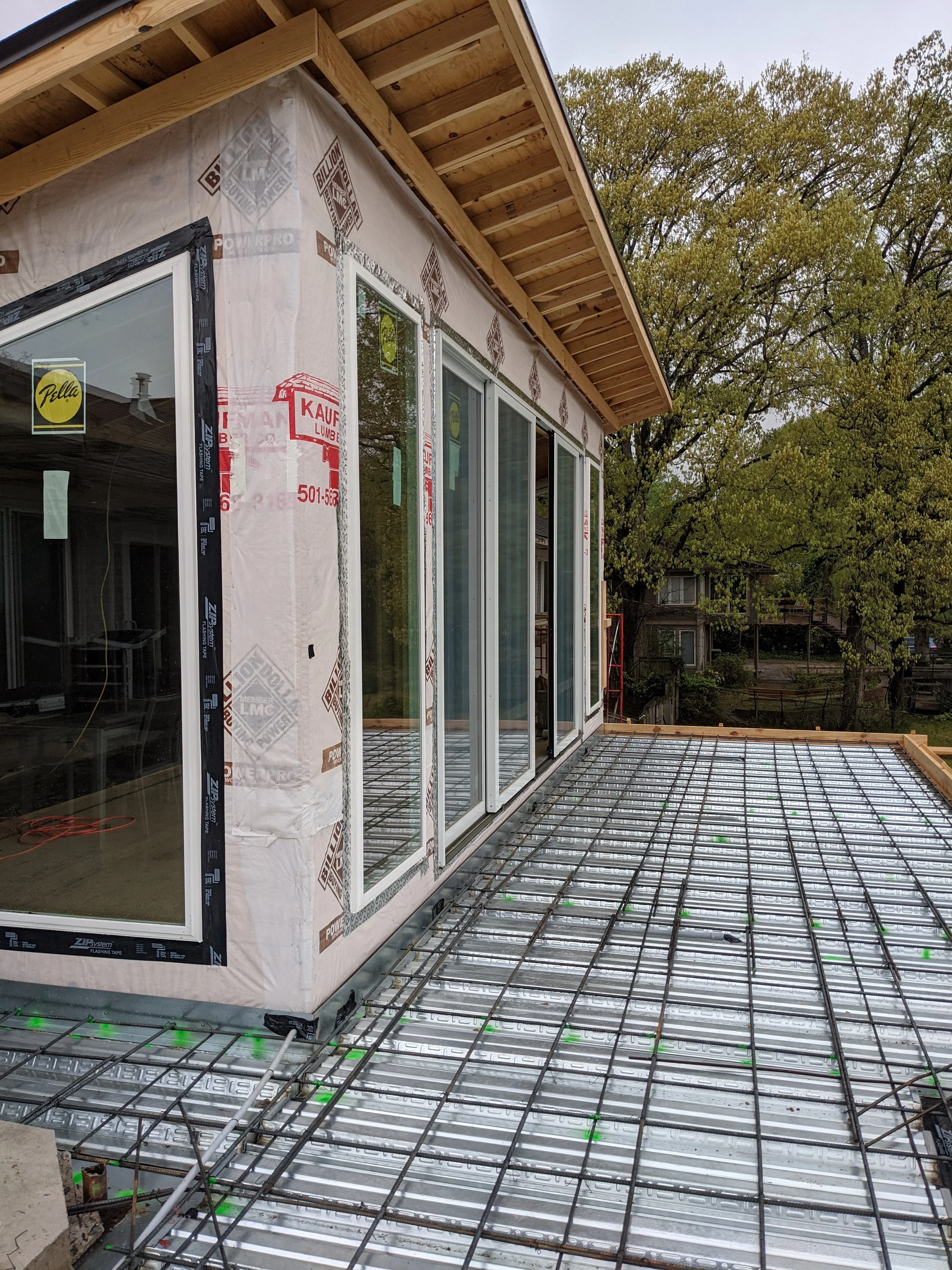 Construction site showing a house with sliding glass doors and exterior insulation panels, a steel rebar grid for a new deck or balcony floor, and a tree in the background.