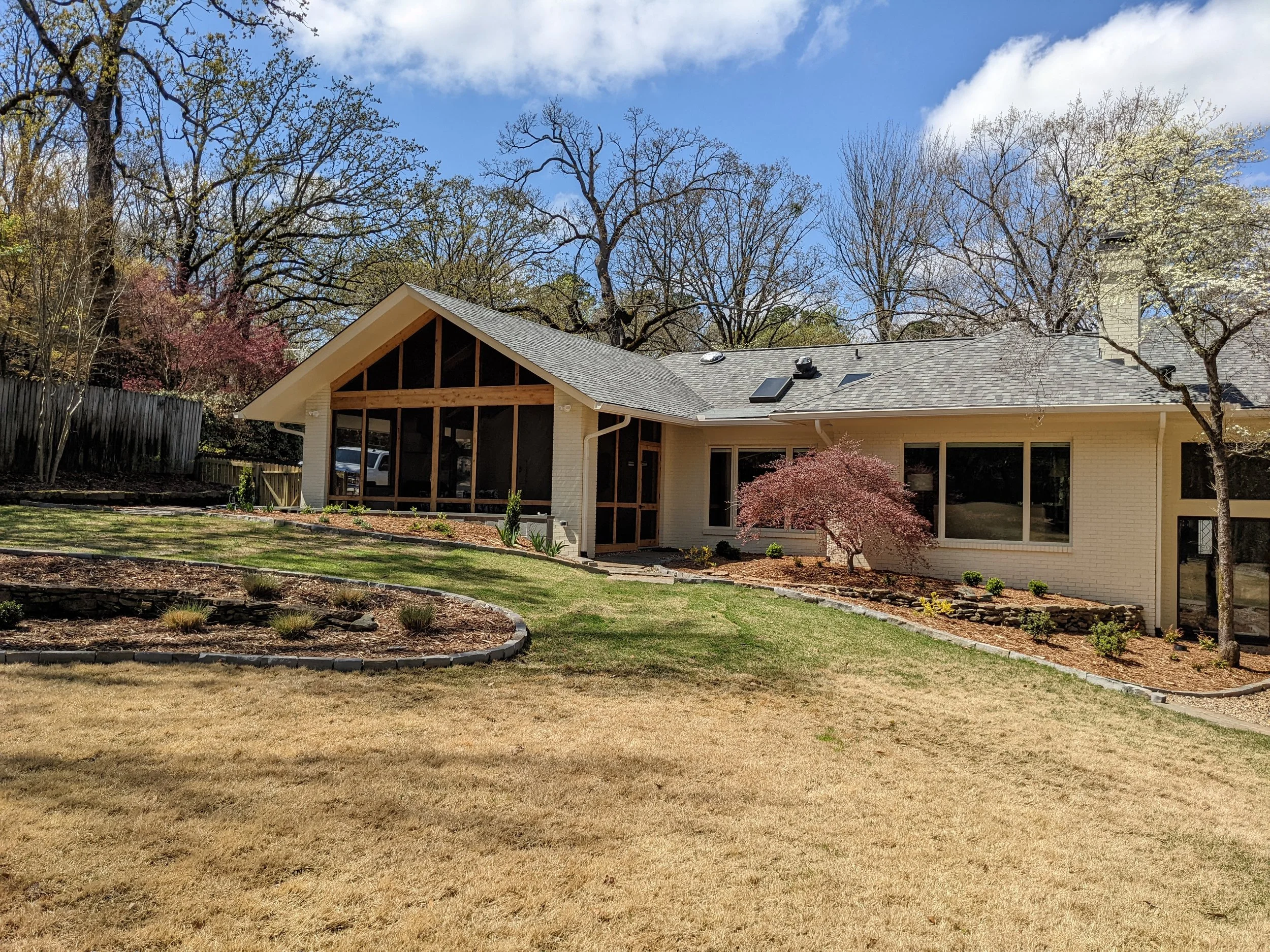 A house with a screened porch, surrounded by trees with budding and blooming branches, a lawn, and a clear blue sky with some clouds.
