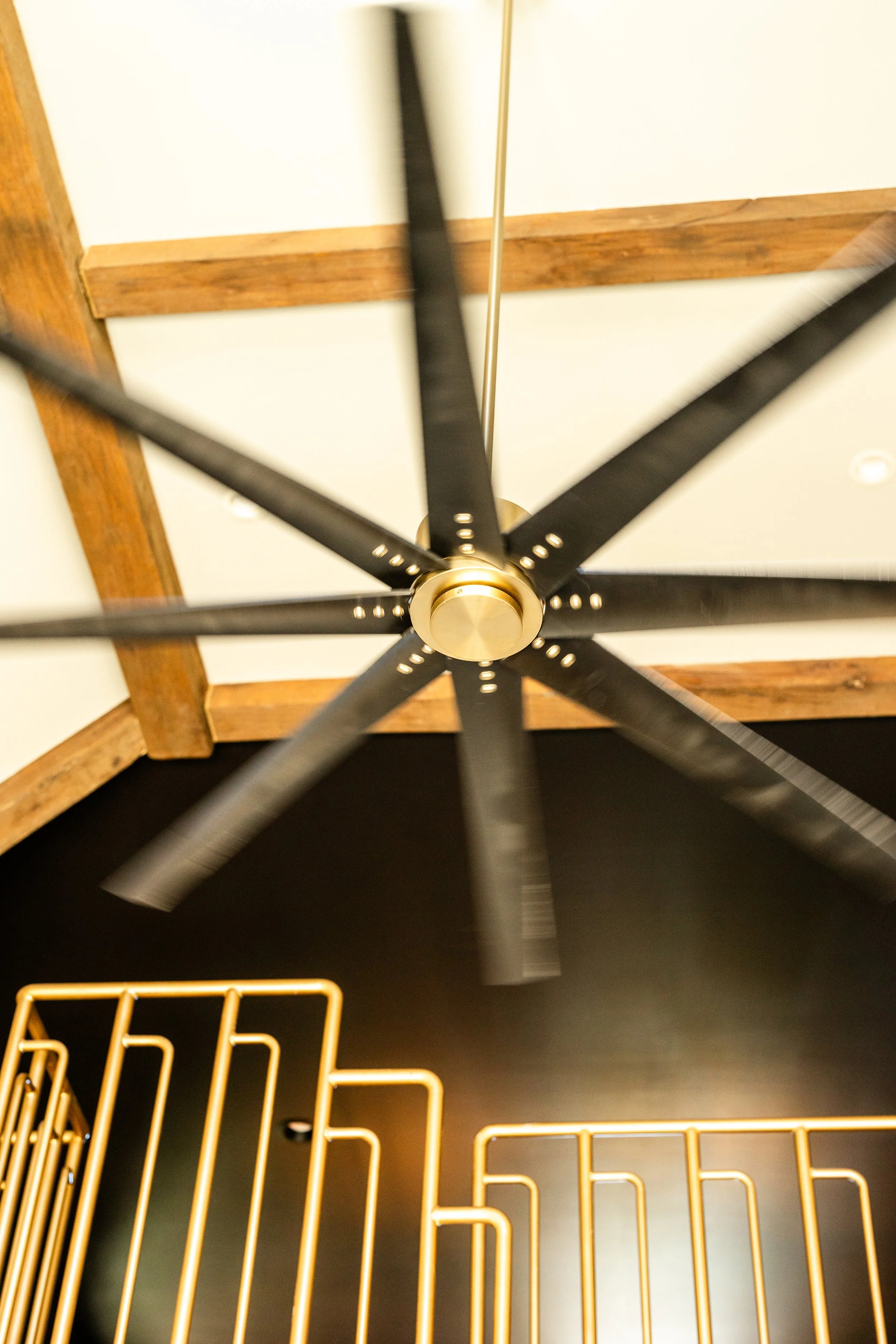 An overhead view of a black ceiling fan with gold accents, mounted on a ceiling with wooden beams and white panels. Below, there is a gold wire basket-style shelf or rack.