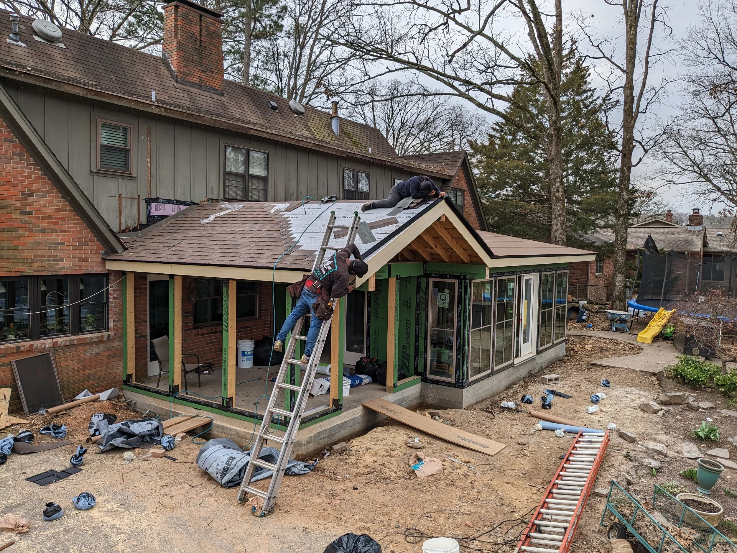 Workers installing a roof on a house extension, with construction materials and tools scattered around a backyard.