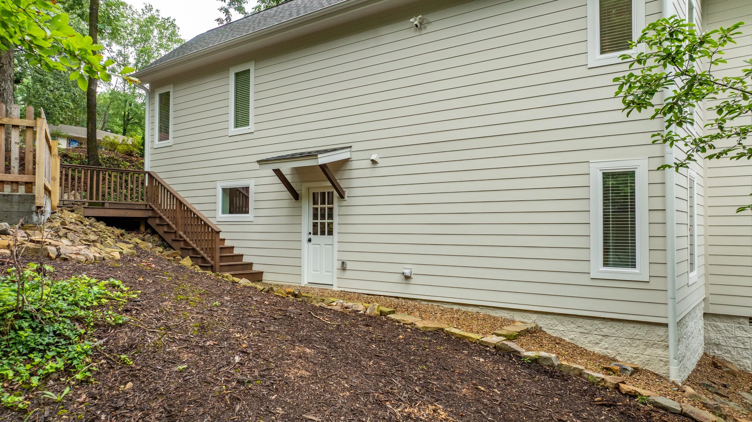 Backyard view of a beige house with vertical vinyl siding, a small staircase with wooden railing leading to a side door, and several windows with blinds. There is a sloped garden area with rocks and dirt, some small plants, and a large tree with gree