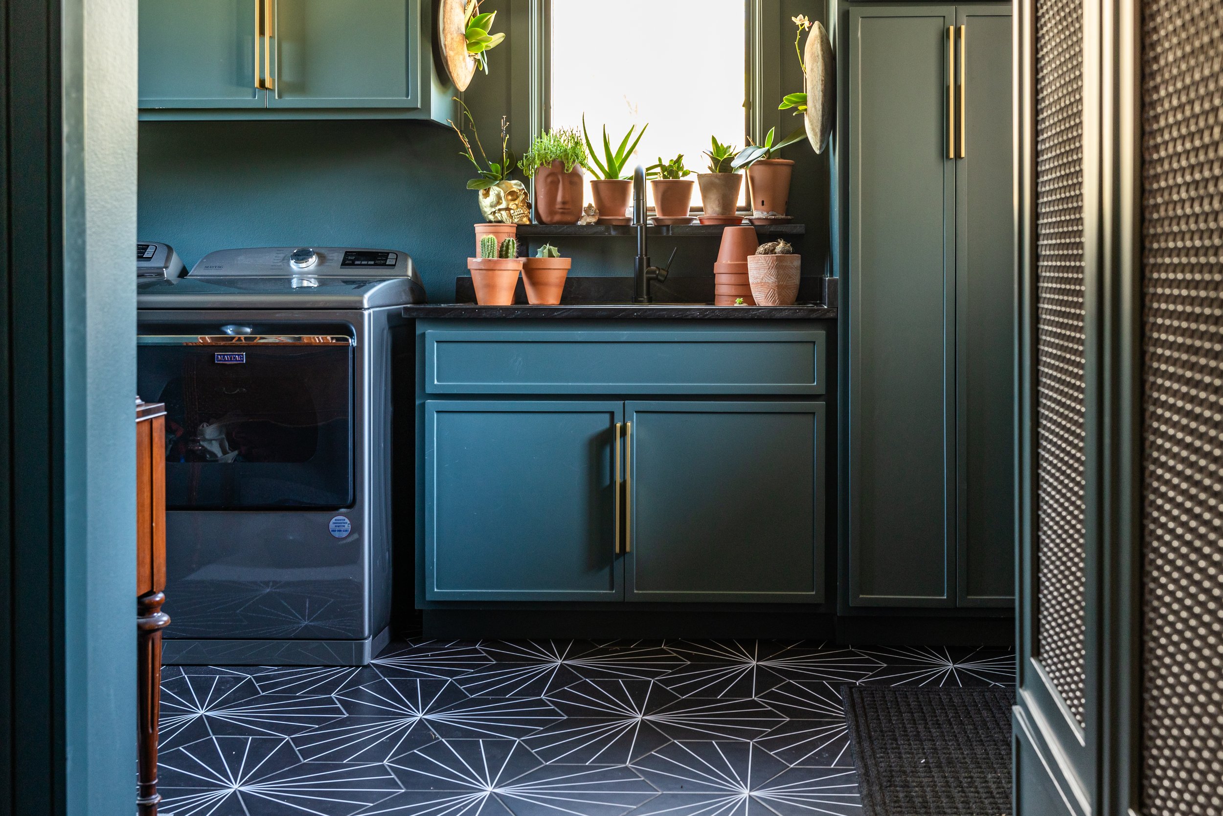 A laundry room with teal cabinets, a black countertop, and potted plants on the windowsill. There is a silver washing machine on the left and a black and white geometric patterned floor. The room is lit by natural light coming through the window.