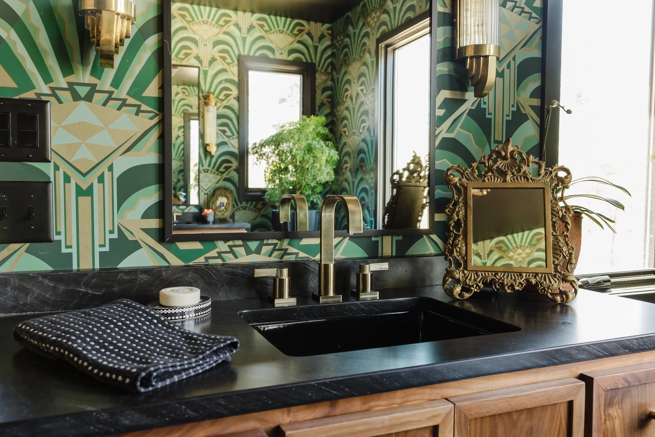 A modern kitchen with a black marble countertop, gold fixtures, a decorative gold picture frame, and a vibrant green geometric patterned wallpaper. A mirror reflects a window and a large potted plant.